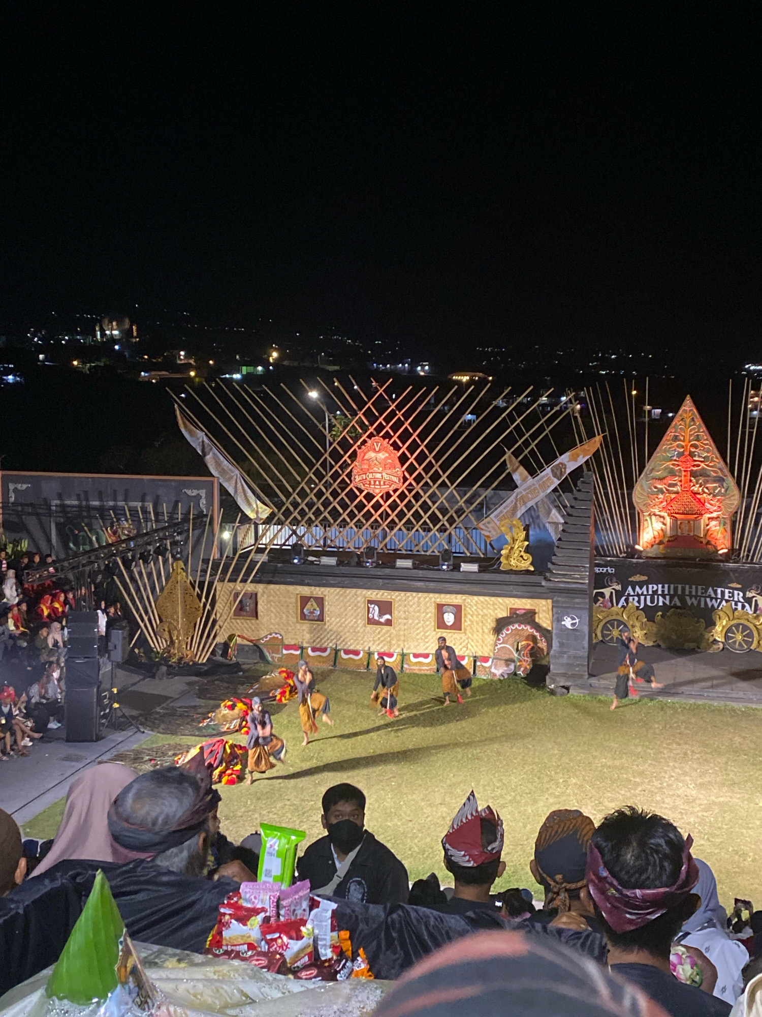 This is a nighttime, low-angle, wide shot of an outdoor amphitheater performance. The stage is decorated with traditional Indonesian motifs, including a large Wayang kulit-style backdrop. Performers in traditional attire are on the grass stage, engaged in a dance or play. In the foreground, a crowd of spectators is seated, facing the stage, with some individuals wearing traditional headdresses. The background shows a city skyline illuminated at night. The overall atmosphere is festive, with a cultural performance taking place.