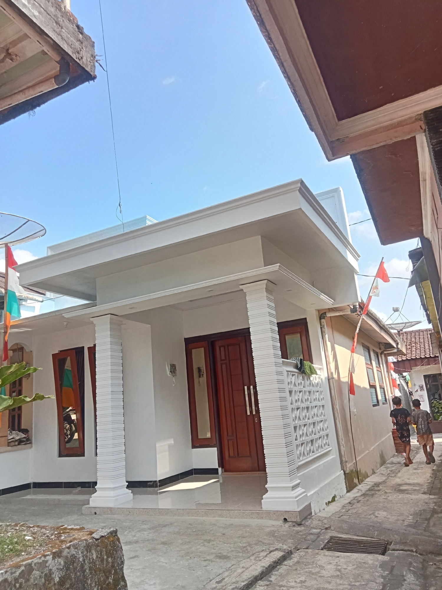 In the city of Pancurwening, Indonesia, a white house with a modern design stands under a clear blue sky. The house features a prominent white-trimmed roof with multiple levels and decorative columns supporting a porch. The entrance has double wooden doors, and windows with wooden frames are visible. Flags, likely representing Indonesia, are attached to the house. A narrow pathway runs alongside the house, paved with concrete and featuring a drainage grate. Two young boys, dressed casually, walk away from the viewer down this pathway. The environment is a residential area, with other buildings and rooflines visible in the background, suggesting a densely populated urban setting. The overall impression is of a sunny day in a tropical locale.