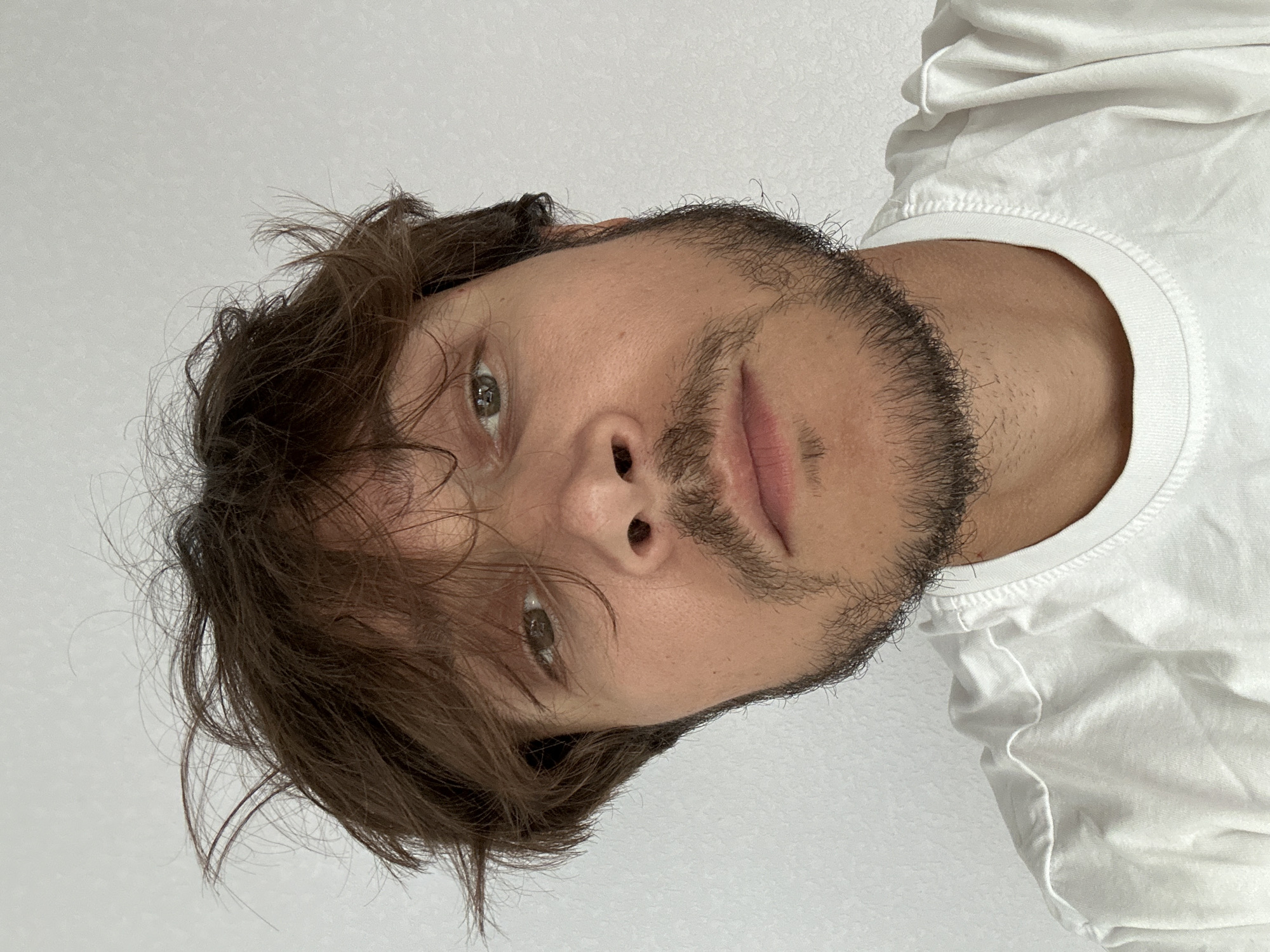 A young man with messy brown hair and a light beard is captured in a close-up, head-and-shoulders selfie against a plain, light-colored background, possibly a wall in an indoor setting. He is wearing a simple white t-shirt. The image is cropped to focus on his face and upper chest, with a slightly tilted perspective that draws the viewer's attention to his features. The lighting appears to be natural, casting soft shadows and highlighting the texture of his skin and facial hair. The overall impression is casual and unposed, suggesting a moment captured in everyday life. There is no information in the image to indicate it is located in Kyiv, Ukraine.