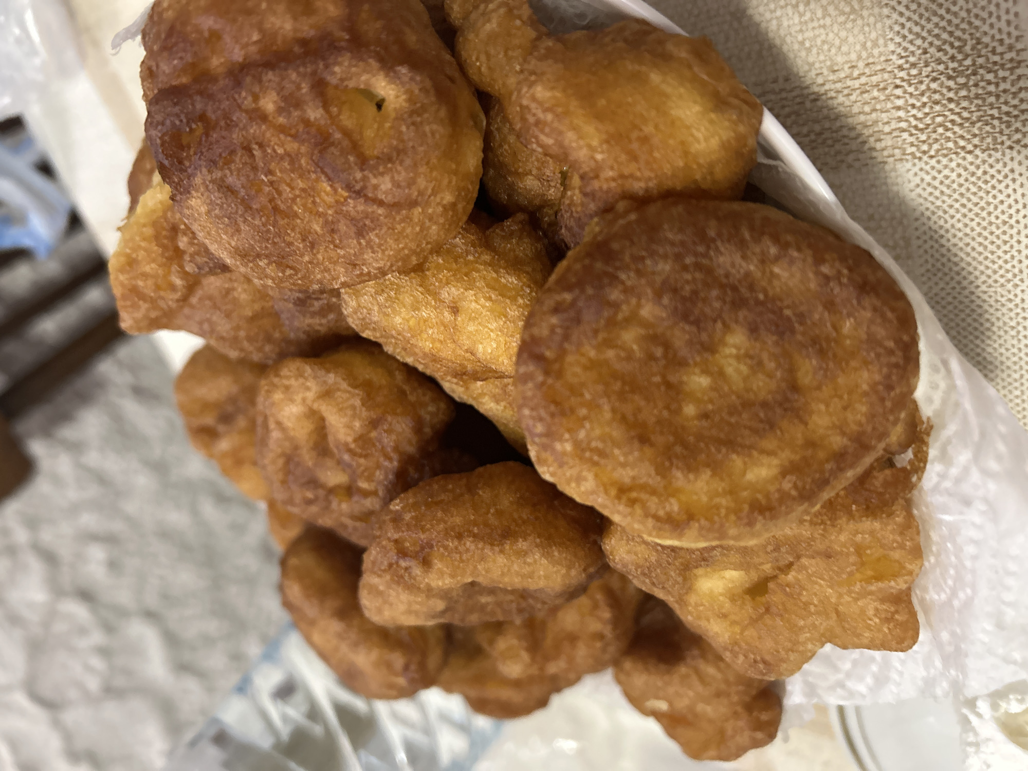 The image shows a close-up view of a pile of golden-brown fried dough pastries. They appear to be freshly cooked and are piled on top of each other in a white container, possibly a bowl or plate. A crumpled white paper towel is visible, likely used to absorb excess oil. The background shows a neutral-colored surface, possibly a tablecloth or counter. There is no indication of a location in Cherven, Bulgaria, or any information about people, actions, or interactions.