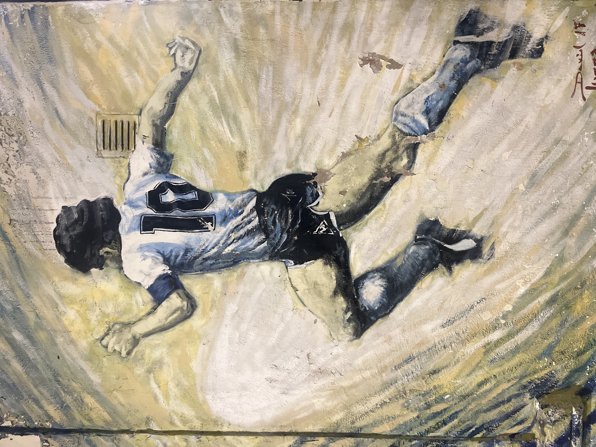 This media file displays a dynamic mural or painting of a male soccer player captured in mid-air. The figure is depicted in a light blue and white striped jersey with the prominent number "10" on his back, black shorts, and blue socks or boots. His body is angled diagonally across the frame, with arms outstretched and one leg bent, conveying a moment of intense athletic action, such as a dive or fall. Given the location context of Buenos Aires, Argentina, and the iconic jersey number, the subject is almost certainly Diego Maradona.
The artwork exhibits a textured, distressed quality, characteristic of a mural, with visible brushstrokes and areas where the paint appears cracked or peeled. The background features abstract, streaking patterns in shades of yellow, beige, and gray, which create a sense of movement radiating from the player. A small, rectangular grate or vent is visible on the upper left side of the background, suggesting an urban setting. On the far right edge, a partially visible, stylized signature or text in a reddish-brown hue is present. The painting effectively captures a powerful and celebrated moment of a sporting legend.