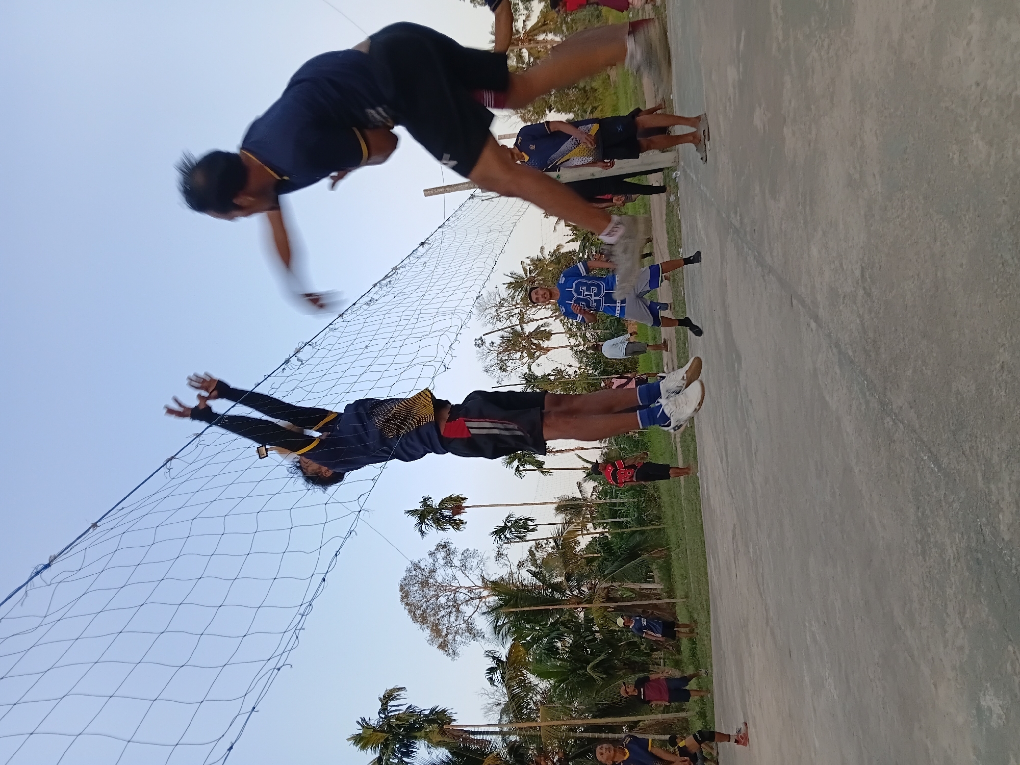 In Margo Rukun, Indonesia, an outdoor volleyball game is underway on a concrete court. Two male players are the central focus, captured mid-action. One player, wearing a dark blue shirt with yellow trim and black shorts, is jumping high to spike the ball, his arms extended above a net. The other player, also in a dark blue shirt and black shorts, is in the foreground, his body blurred due to motion as he appears to be mid-hit or follow-through, jumping with his legs extended. Several other individuals, men and women, are visible in the background, some observing the game and others standing on the court. The environment is lush with palm trees and other tropical vegetation surrounding the court, suggesting a rural or semi-rural setting. The sky is clear and pale blue, indicating daytime. The scene captures a dynamic moment of athletic competition within a community gathering.