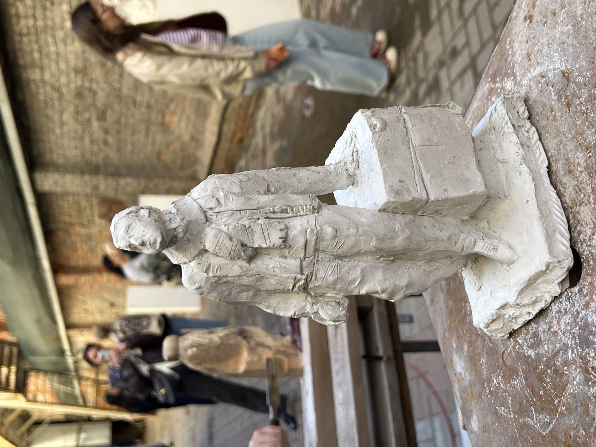 The image shows a white, sculpted figure of a man holding a box, likely a suitcase, in an indoor space. The sculpture is set on a textured, possibly metallic surface.
In the background, blurred figures are visible. A woman is visible in the upper part of the image, wearing a light jacket and blue jeans. A structure of scaffolding can be seen behind the artwork.
The image's style is amateur photography with a shallow depth of field.