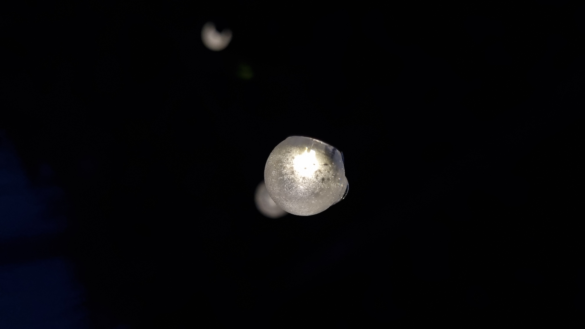 A close-up, low-light shot shows a string of round, glowing lights against a dark background. The light in the foreground is the most in focus, appearing as a translucent sphere with a bright light source within. A liquid, possibly condensation, can be seen on its surface. Two other lights are visible in the background, further out of focus and appearing as soft, circular blurs. A hint of green foliage is visible in the upper right quadrant, suggesting an outdoor setting. The image captures a festive or decorative illumination. The location is Vysokoe, Russia.