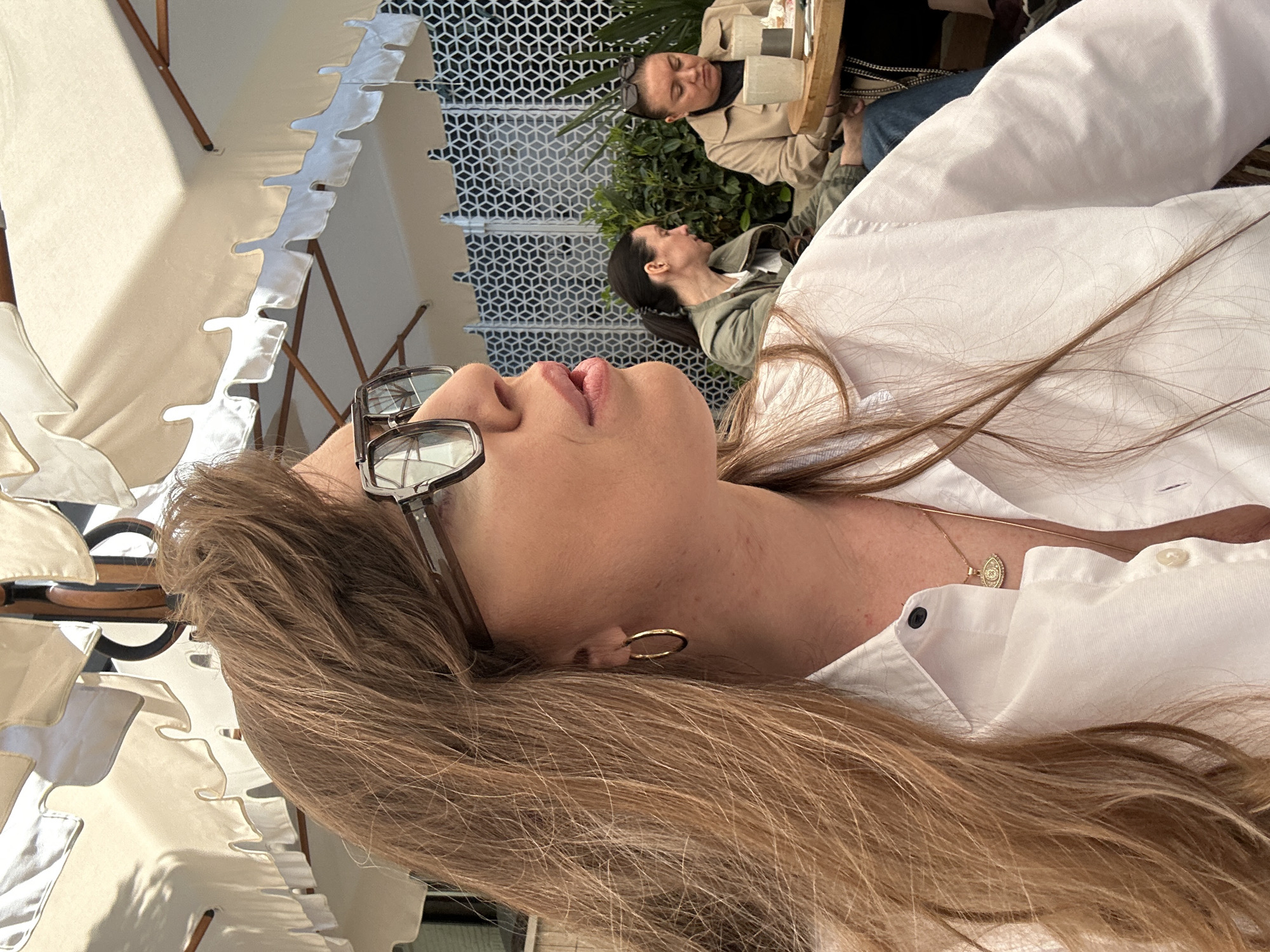 An overhead shot captures a moment at an outdoor cafe or restaurant patio in Tbilisi, Georgia, on a bright, sunny day. The image is oriented sideways, but viewed correctly, it shows a close-up of a woman in the foreground, with her head tilted back, looking upwards. Only her chin, mouth, nose, and forehead are visible, framed by black-rimmed glasses pushed onto her nose. Her long, light brown hair cascades over her shoulders. She wears a white collared shirt, a gold hoop earring in her visible ear, and a delicate gold necklace with a small oval medallion.
In the mid-ground, two other women are seated at a wooden table, partially obscured by the foreground subject's hair. One woman, with dark hair, wears a green hooded jacket and looks towards her companion. The second woman, wearing a beige trench-style coat with sunglasses perched on her head, also appears to be engaged in conversation or observing something on the table, where a cup is visible.
The background features decorative white canvas patio umbrellas, casting shadows, and a modern, geometric patterned metal screen or wall, behind which some green foliage is visible, likely potted plants or bushes. The strong sunlight and clear shadows indicate it is daytime. No visible text is present in the image.
