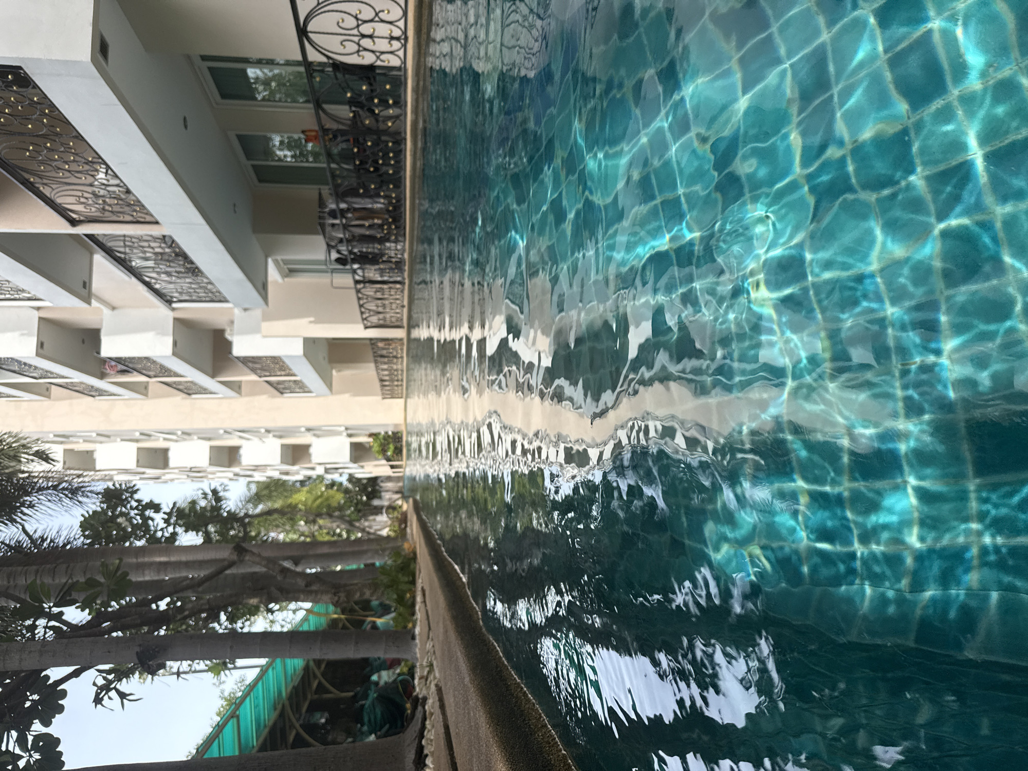 An angled, ground-level shot looks across a turquoise swimming pool at a modern, multi-story building with balconies. The pool tiles are visible through the rippling water, reflecting the building's facade and the sky. Palm trees and a covered green awning are visible to the left of the pool, framing the building. The building appears to be a hotel or residential complex in Pattaya City, Thailand, given its architectural style and tropical vegetation. No people are visible in the image.