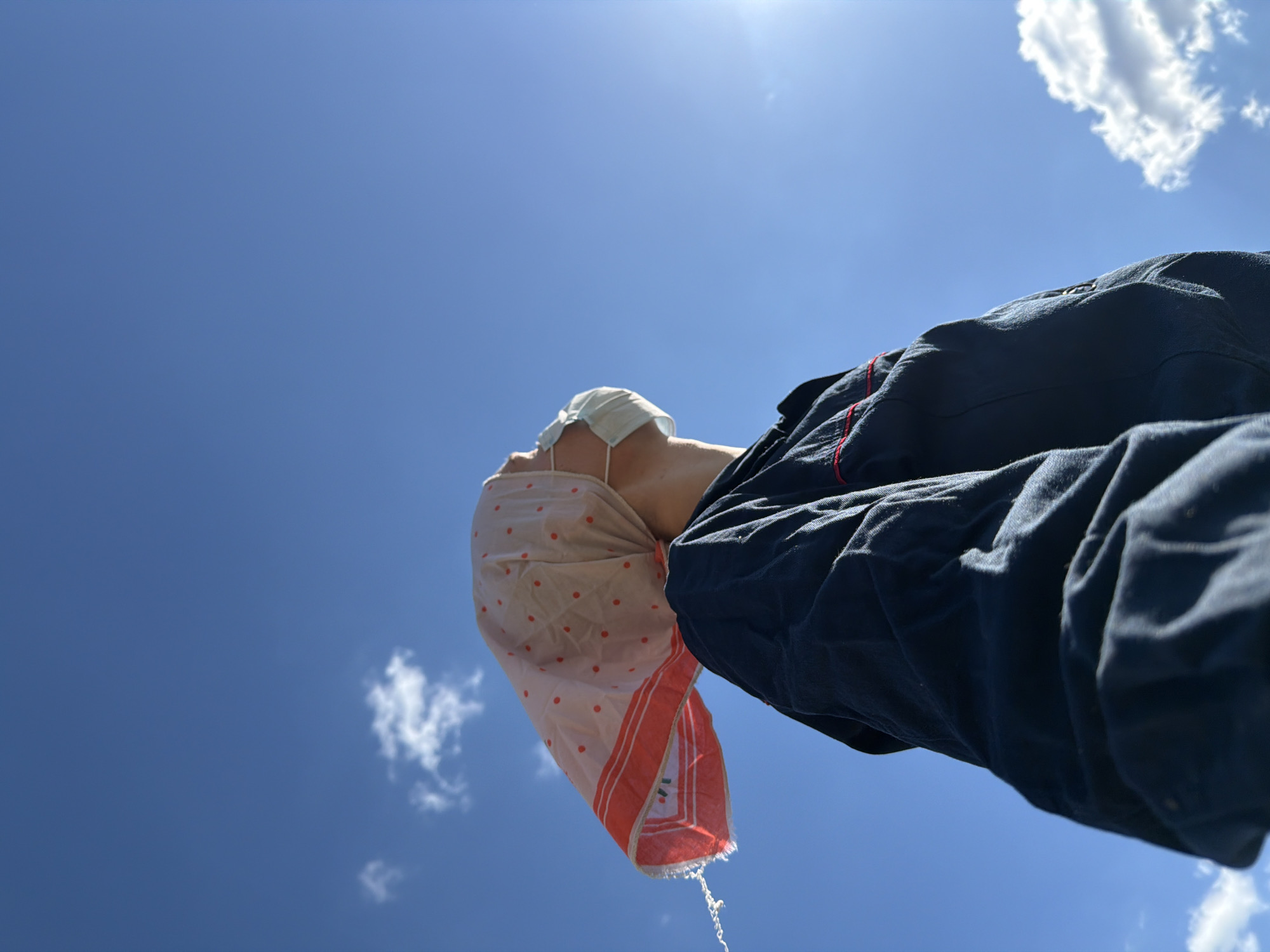 A person wearing a medical mask, a pink and white polka-dot scarf, and a dark blue jacket is seen from a low angle against a clear blue sky with scattered white clouds. The person is looking upward, and what appears to be water is dripping from the scarf. The location is assumed to be Chaikovsky, Russia.