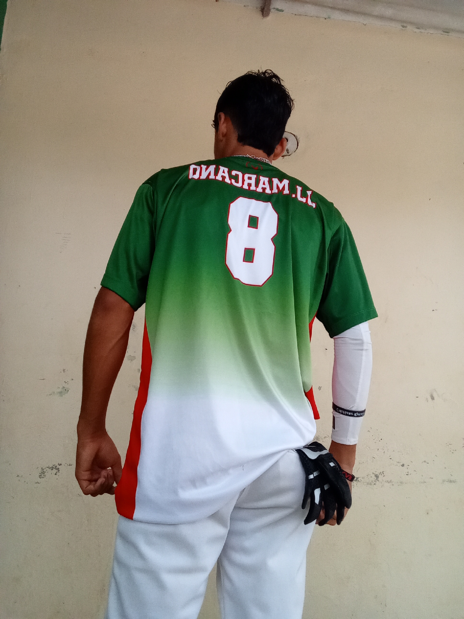 A person wearing a green, white, and red baseball jersey with the number 8 and the name "MARCANO" on the back stands with their back to the camera. They are also wearing white pants and a black and white baseball glove. The person is standing against a light-colored wall in what appears to be an outdoor area, possibly in the city of Juan Griego, Venezuela.
