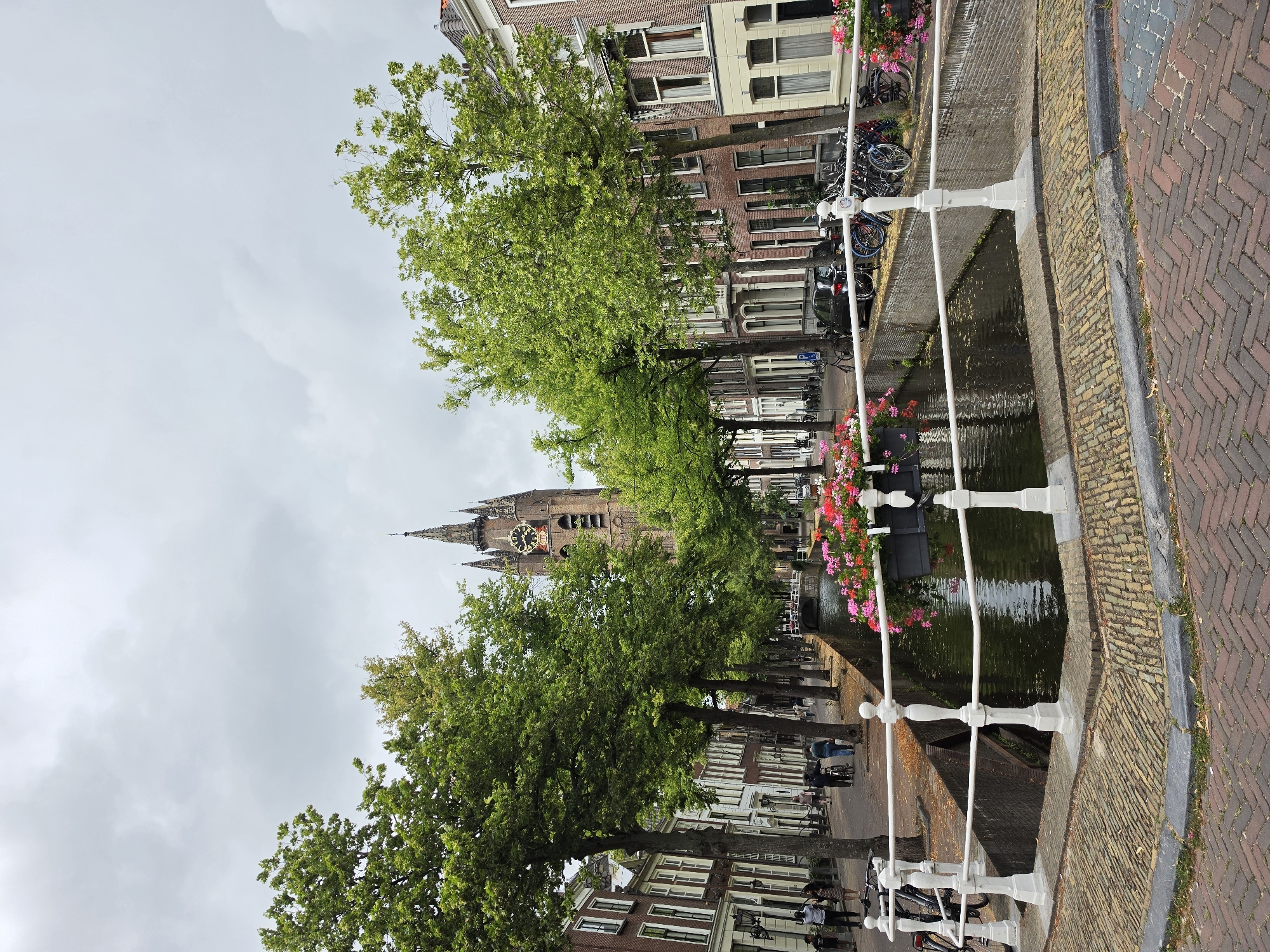 The image depicts a canal scene in Delft, Netherlands, with a prominent tower in the background. The canal is lined with buildings on both sides, featuring traditional Dutch architecture with brick facades and white-trimmed windows. Lush green trees line the canal, casting shadows on the brick-paved street and sidewalk. A white railing with decorative posts borders the canal. Several people are visible walking along the sidewalk and near bicycles parked along the canal. The canal itself contains water with reflections of the surrounding environment. Flowers in planters add a splash of color to the scene, positioned on the edge of the canal. A bridge crosses the canal in the distance. The sky is overcast, suggesting a cloudy day. The iconic Nieuwe Kerk, or New Church, with its clock tower, is identifiable as the landmark in the background, confirming the location as Delft.
