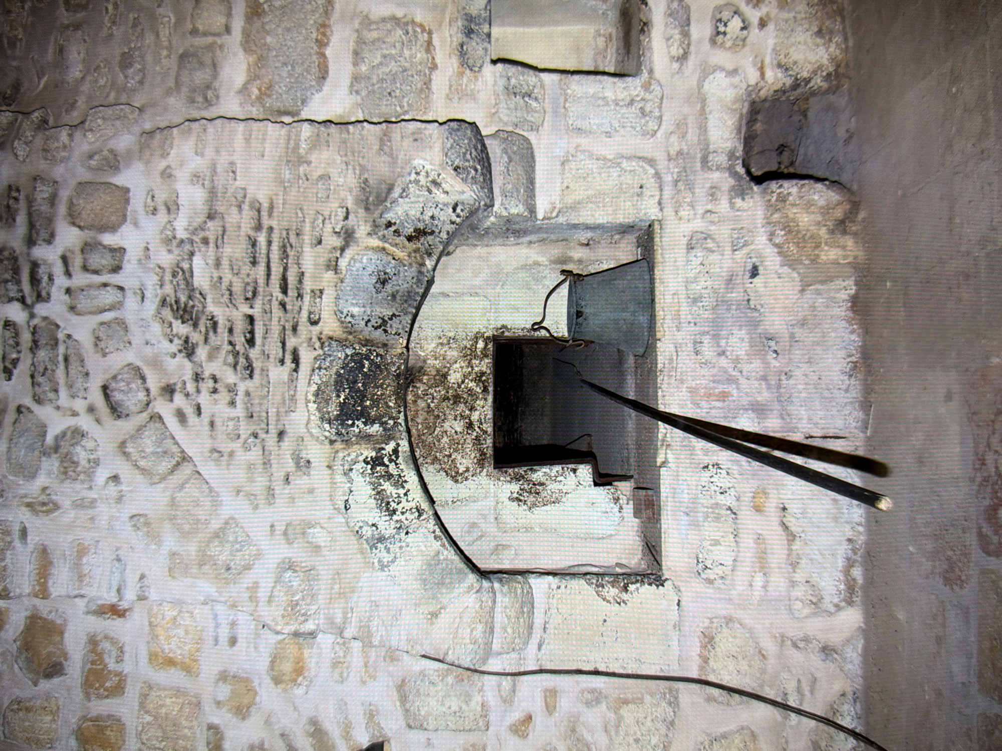 The image shows a close-up view of a stone oven or hearth. The structure is made of rough-hewn stones of various sizes and colors, mortared together. The opening of the hearth is arched and appears to be blackened with soot and residue, suggesting past use. Inside the opening, a dark space is visible. To the right of the opening, a metal bucket with a handle rests on a stone ledge. Two long, dark poles, possibly fire-tending tools, are angled across the lower part of the hearth opening. A dark, thin cable runs along the left side of the image, adjacent to the stone wall. The surrounding area appears to be a floor made of concrete or packed earth. The image is a static, medium shot of this architectural feature. The location is not identifiable from the visual information provided.