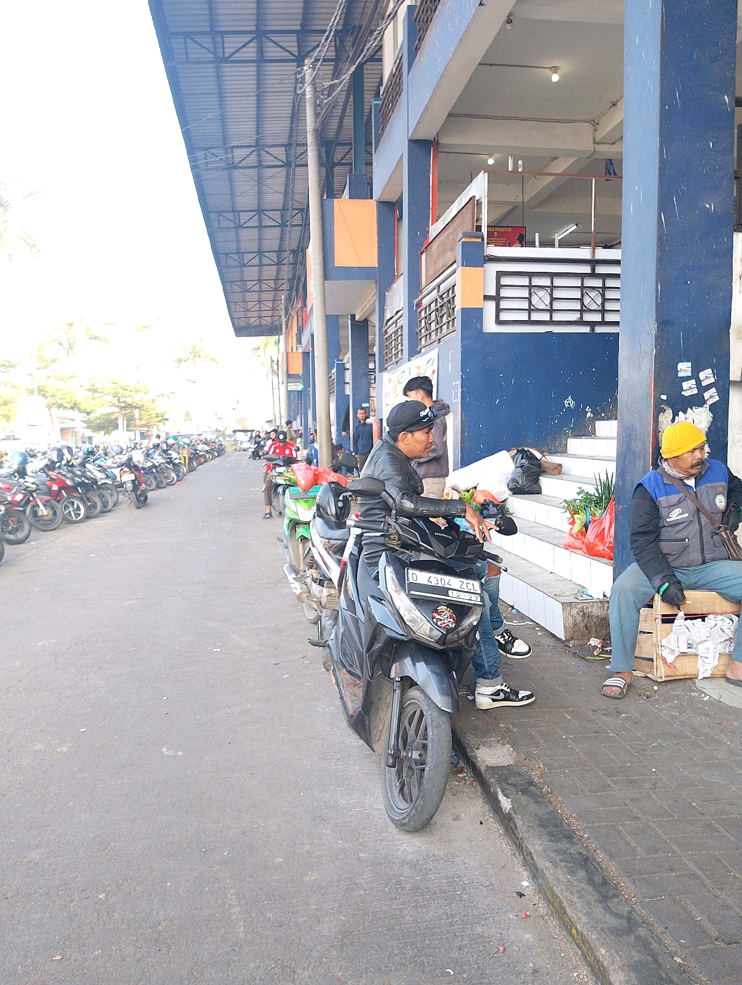 The image depicts a street scene in Lembang, Indonesia, likely a marketplace or parking area, with a row of parked motorcycles on the left side of the frame. The architecture suggests a multi-story building with blue and orange accents. On the right side of the street, two men are present. One man, wearing a yellow beanie, is seated on a wooden crate, dressed in a grey vest with blue trim and grey pants. He appears to be engaged in selling goods, with a selection of items, including red plastic bags filled with produce and a black trash bag, visible on the steps behind him. Another man, wearing a black leather jacket and a black cap, is seated on a black motorcycle, facing towards the viewer and interacting with the seated man. The motorcycle has a license plate that reads "D 4304 ZCL". The ground is paved with asphalt and cobblestones, and there is some debris on the street and sidewalk, indicating a busy public area. The sky is bright, suggesting daylight, and trees are visible in the background on the left.