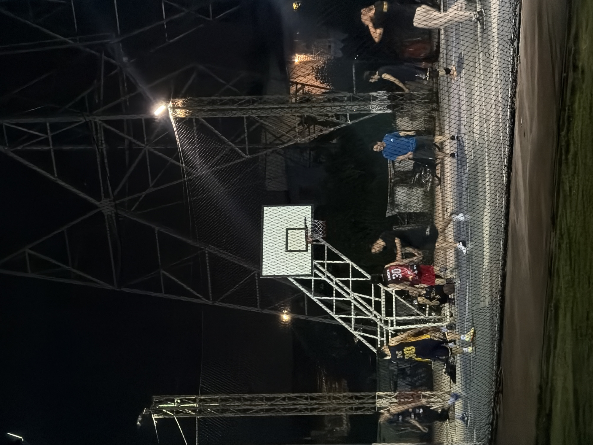 The image shows an outdoor basketball court at night, possibly located in Chang Phueak, Thailand. The court is surrounded by a tall chain-link fence. Several people are present on and around the court, some wearing basketball attire and some observing. A basketball hoop and backboard are visible, illuminated by artificial lighting. The environment appears to be urban, with buildings or structures visible in the background beyond the fence. There is also a patch of what appears to be grass on the right side of the image.