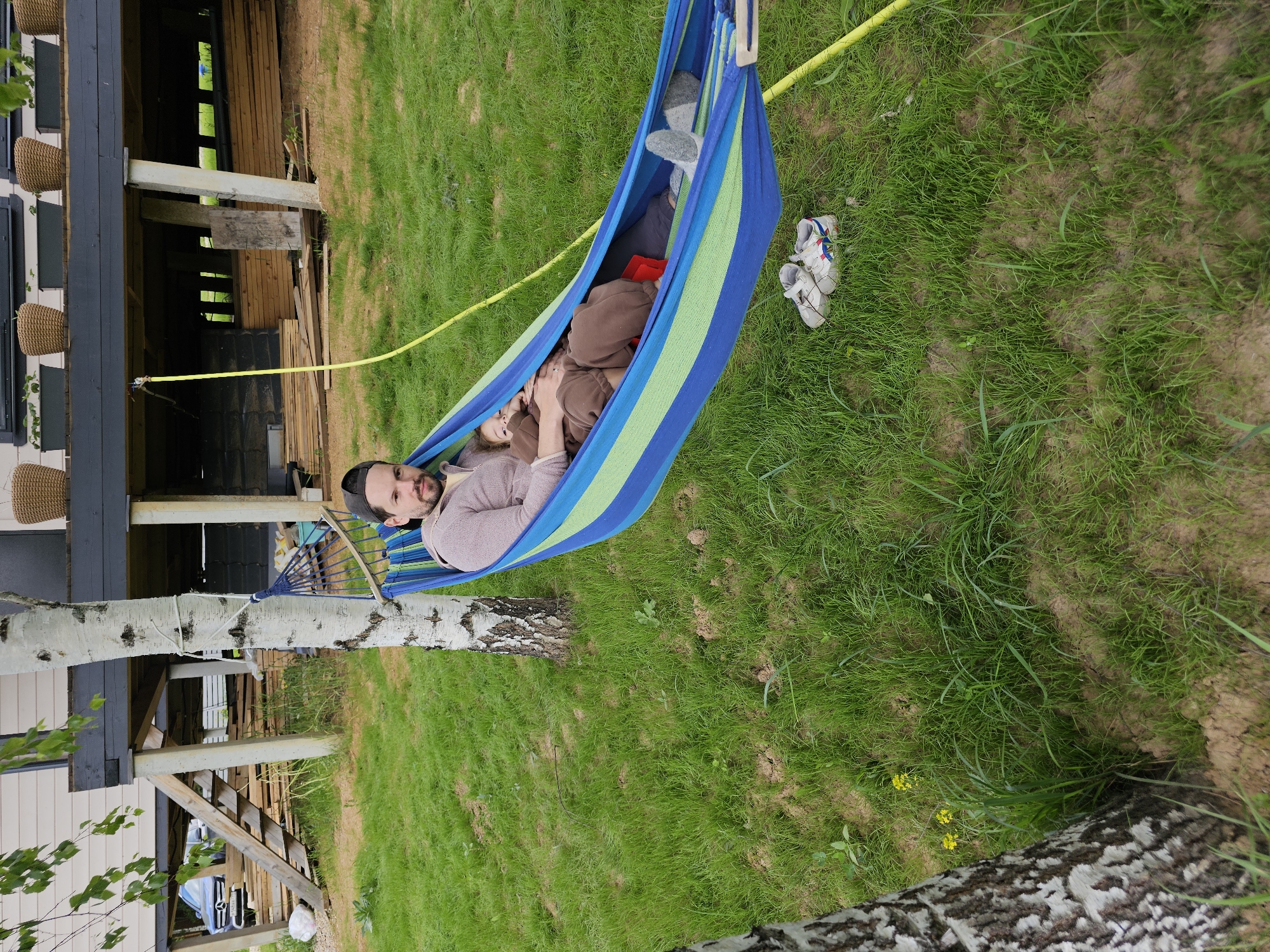 A man and child are lying in a striped blue and green hammock suspended between a birch tree and the support structure of a building. The man is wearing a beanie and light-colored sweater. The child, mostly obscured, is wearing brown clothing. A pair of white athletic shoes is on the grass near the hammock. A yellow rope is present. The scene appears to be in a yard with green grass. Мышкино, Russia is not discernable from the image.