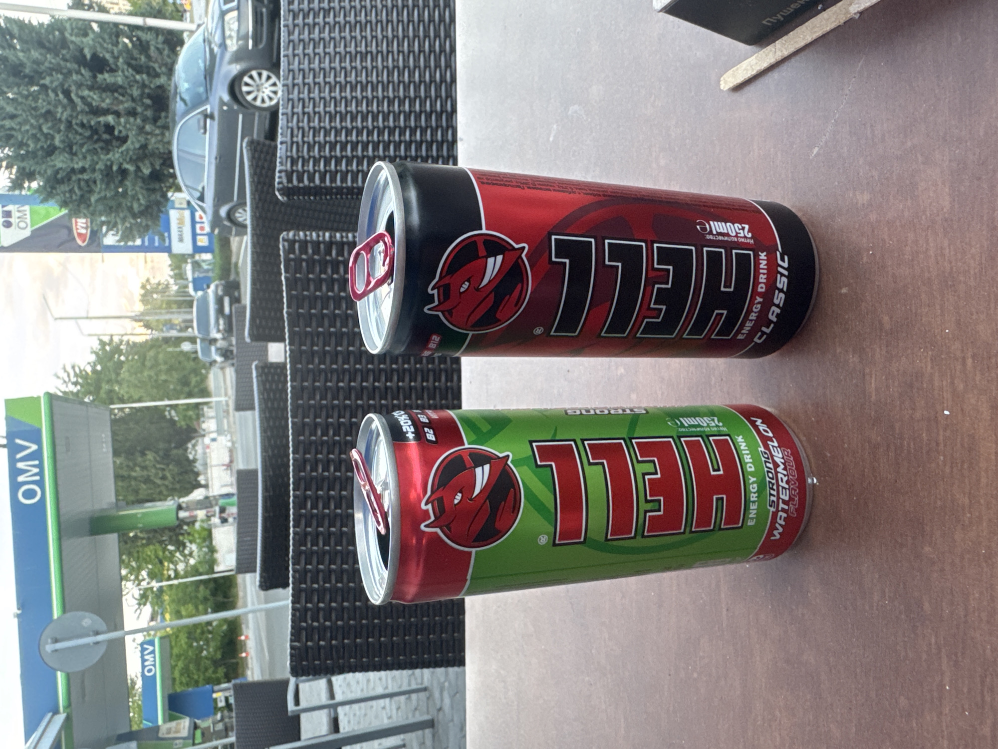 The image shows two cans of Hell energy drink sitting on a brown table, one red and black, the other green and red. A wicker chair can be seen behind the cans. In the background, an OMV gas station is visible on the left side of the frame. A parked car is visible in the background, alongside trees and a road. Based on this information, the location is likely a table at an establishment near an OMV gas station in Stara Zagora, Bulgaria.