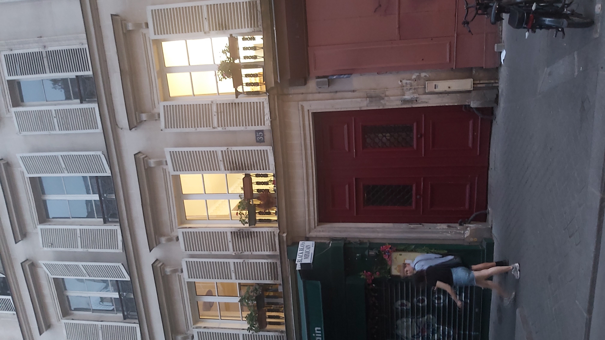 The image captures a street scene in Paris, France, focusing on the exterior of a building with multiple windows and a large red double door. The building's facade is light-colored, and the windows are adorned with white shutters, many of which are open. Some windows feature flower boxes with plants. The number "35" is visible on the building. To the left, a green awning for a shop labeled "ain" is partially visible, with a sign above it reading "BLACK BEANS MEXICAN." A closed metal shutter is seen below the awning. In front of the building, two individuals are walking on a paved sidewalk. One person, wearing a black t-shirt and denim shorts, is in the foreground. Behind them, another person in a light blue shirt and dark shorts is visible. A dark red double door is the prominent feature on the ground floor. To the right of the door, a pinkish-red section of another building or shop front is visible, along with a bicycle parked on the sidewalk. The overall scene suggests a quiet street in a European city, possibly during the late afternoon or early evening given the lighting.