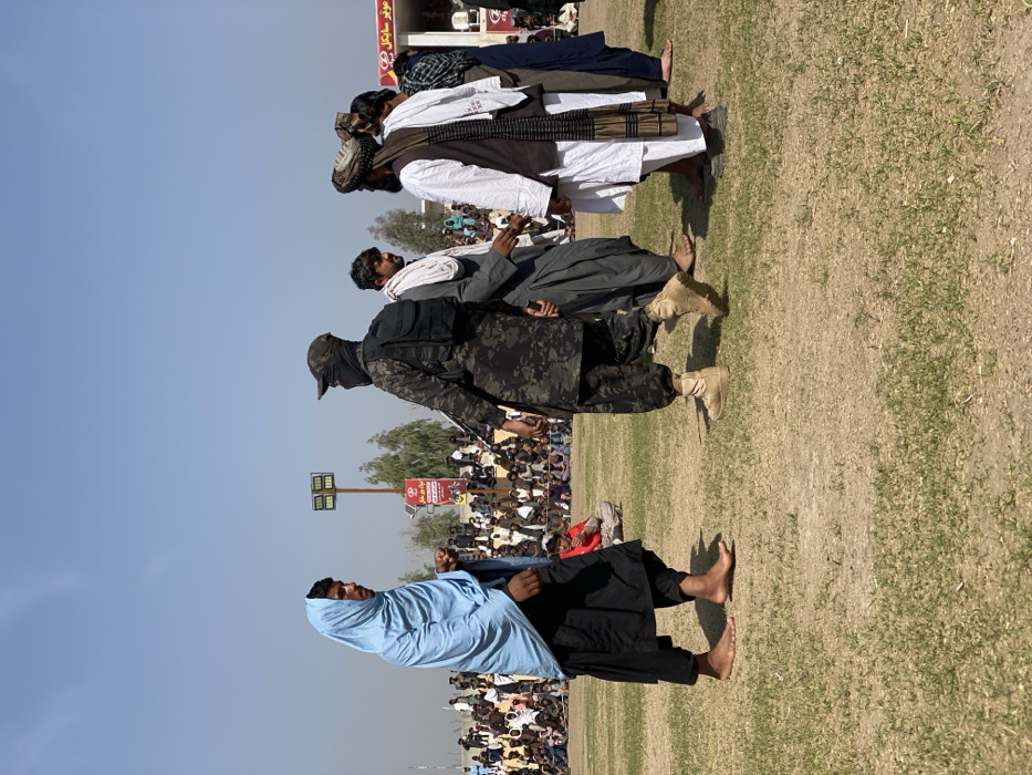 A group of men stand in a line on a grassy field, facing an audience. The men wear traditional clothing, including robes and head coverings. One man wears a camouflage uniform and a face covering. The audience is seated and appears to be watching the men. A light pole is visible in the background. The sky is blue.