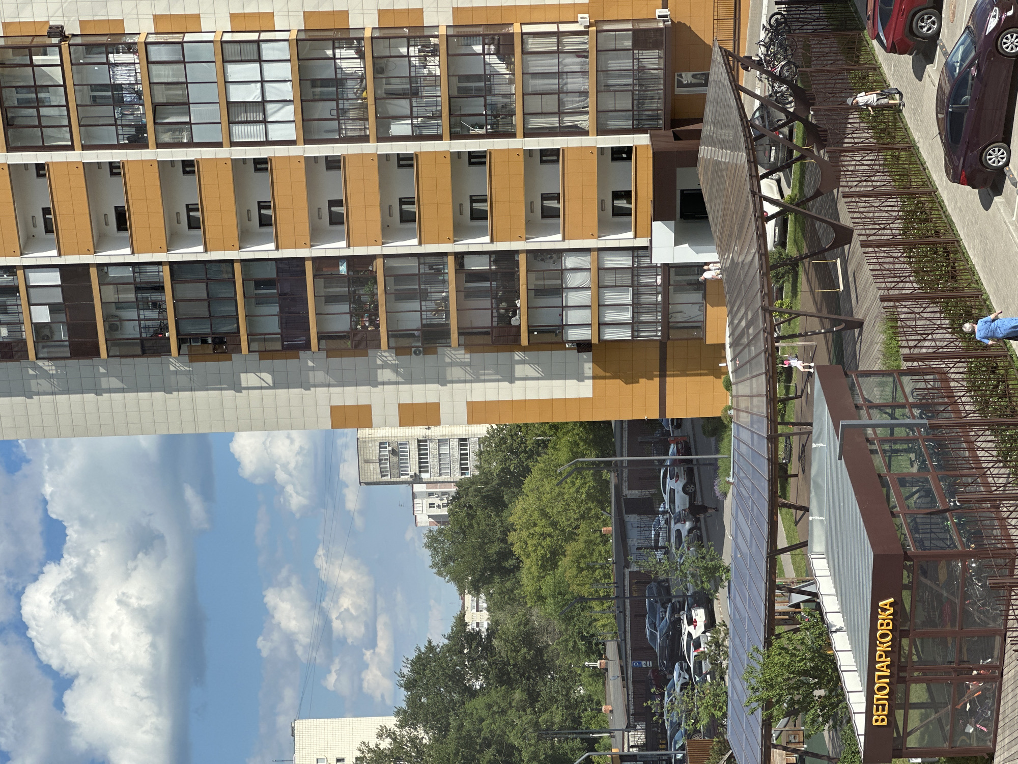 This image shows a modern residential building in Kazan, Russia, with a clear sky and white clouds above. The building features a prominent orange and white facade with numerous balconies and windows. In the foreground, there is a covered bicycle parking area labeled "ВЕЛОПАРКОВКА" (Veloparkovka, meaning bicycle parking in Russian). Several parked cars are visible in a lot next to the building, and a few people are present in the scene, including one person walking on the sidewalk and another near the bicycle parking. The environment appears to be a residential urban area with trees and other buildings in the background.