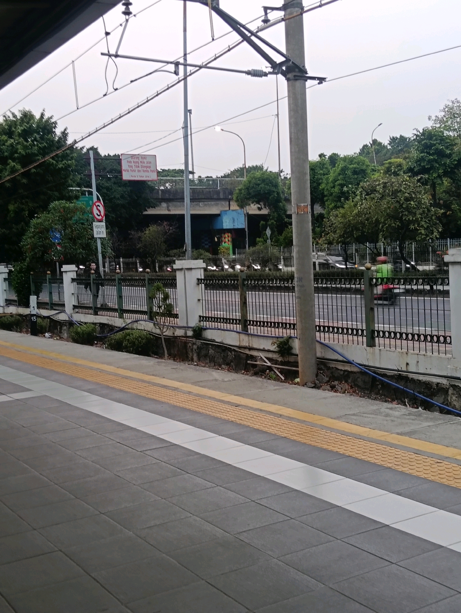 An exterior, medium-shot image captures a view from a train station platform in East Jakarta, Indonesia, looking towards a street and a pedestrian overpass. The platform, paved with grey tiles and marked with tactile paving and white lines, occupies the foreground. To the left, a dark overhang covers part of the scene.
Beyond the platform, a black metal fence with white concrete pillars runs horizontally, separating the station from a busy road. Several vehicles, including cars and a motorcycle with a rider wearing a green jacket, are visible on the road. Trees and foliage line the background, with a multi-level overpass dominating the mid-ground, its concrete structure a muted blue and grey. Overhead, a complex network of electrical wires and support poles suggests the presence of a railway system.
Two traffic signs are visible: a circular red sign with a diagonal bar over a black 'S' indicates no parking, and a rectangular white sign with red text appears to be a warning or regulation notice, though the text is illegible. A lone person, seen from the side and facing away from the camera, stands by the fence, observing the street. The overall atmosphere is overcast, with diffused light suggesting an early morning or late afternoon setting. The scene is set in an urban environment, likely a transit hub.