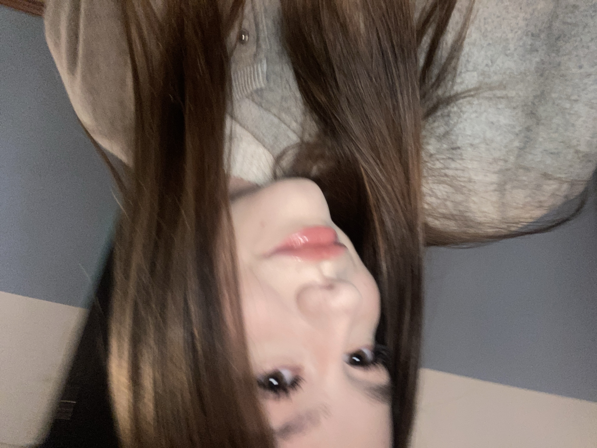 A young woman with long brown hair is the central focus of the image. She is wearing a light-colored cardigan. The background features a wall with a two-toned color scheme, likely in an interior space. The image is taken from a slightly high angle, and the woman's face is turned towards the camera. Location of the photo in Chaikovsky, Russia cannot be determined.