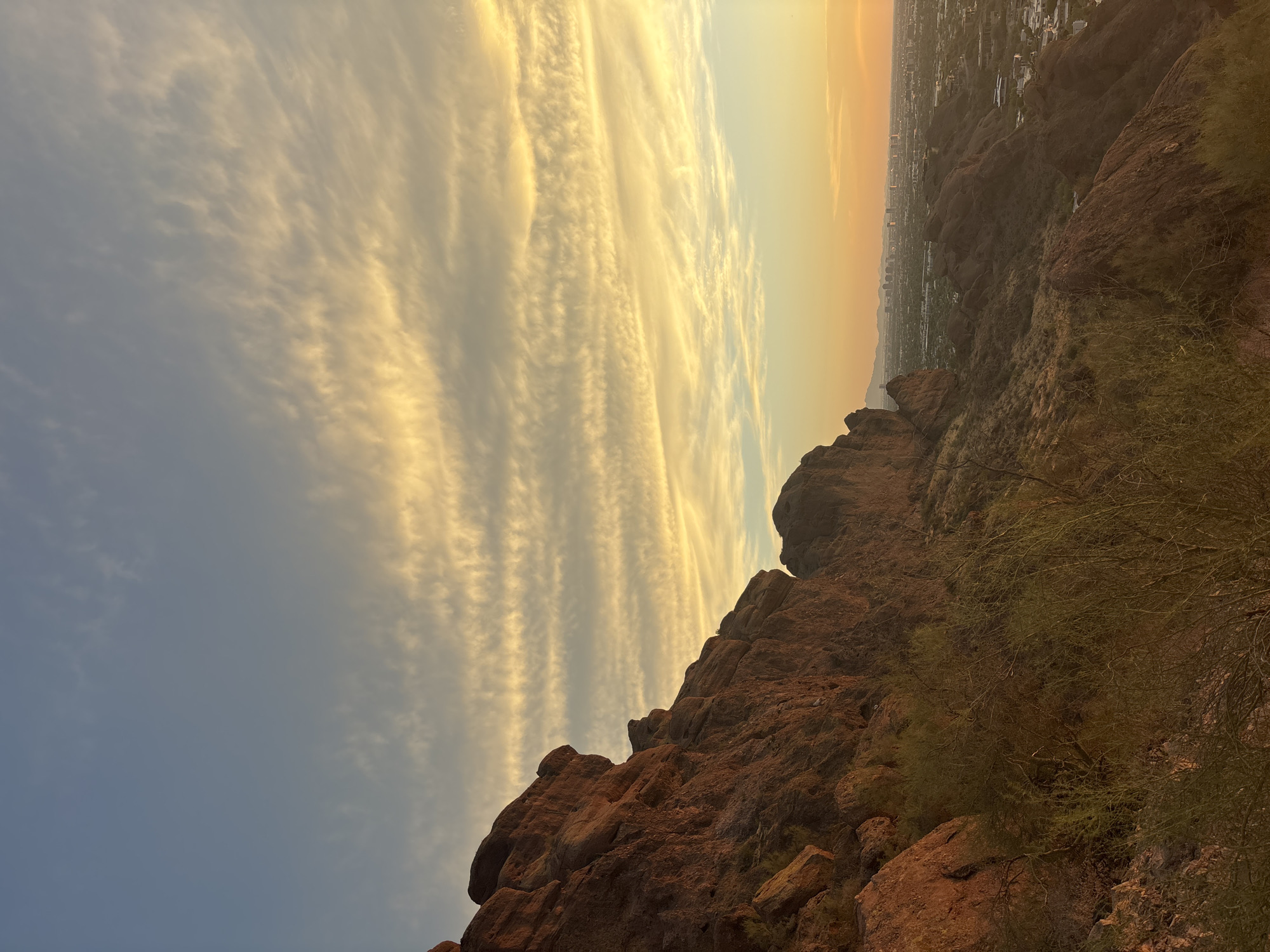 The image depicts a scenic view from the top of a mountain in Phoenix, Arizona, USA. The foreground is dominated by rugged, reddish-brown rock formations and sparse desert vegetation. The camera is angled downwards, looking out over a vast urban landscape that stretches to the horizon. The city is characterized by numerous buildings and structures, suggesting a densely populated area. In the distance, a faint outline of mountains can be seen. The sky above is a blend of soft blues and yellows, with wispy, elongated clouds painted in warm hues of gold and white, indicative of a sunset or sunrise. The lighting suggests either early morning or late evening, casting a warm glow over the scene. There are no people or animals visible in the frame.