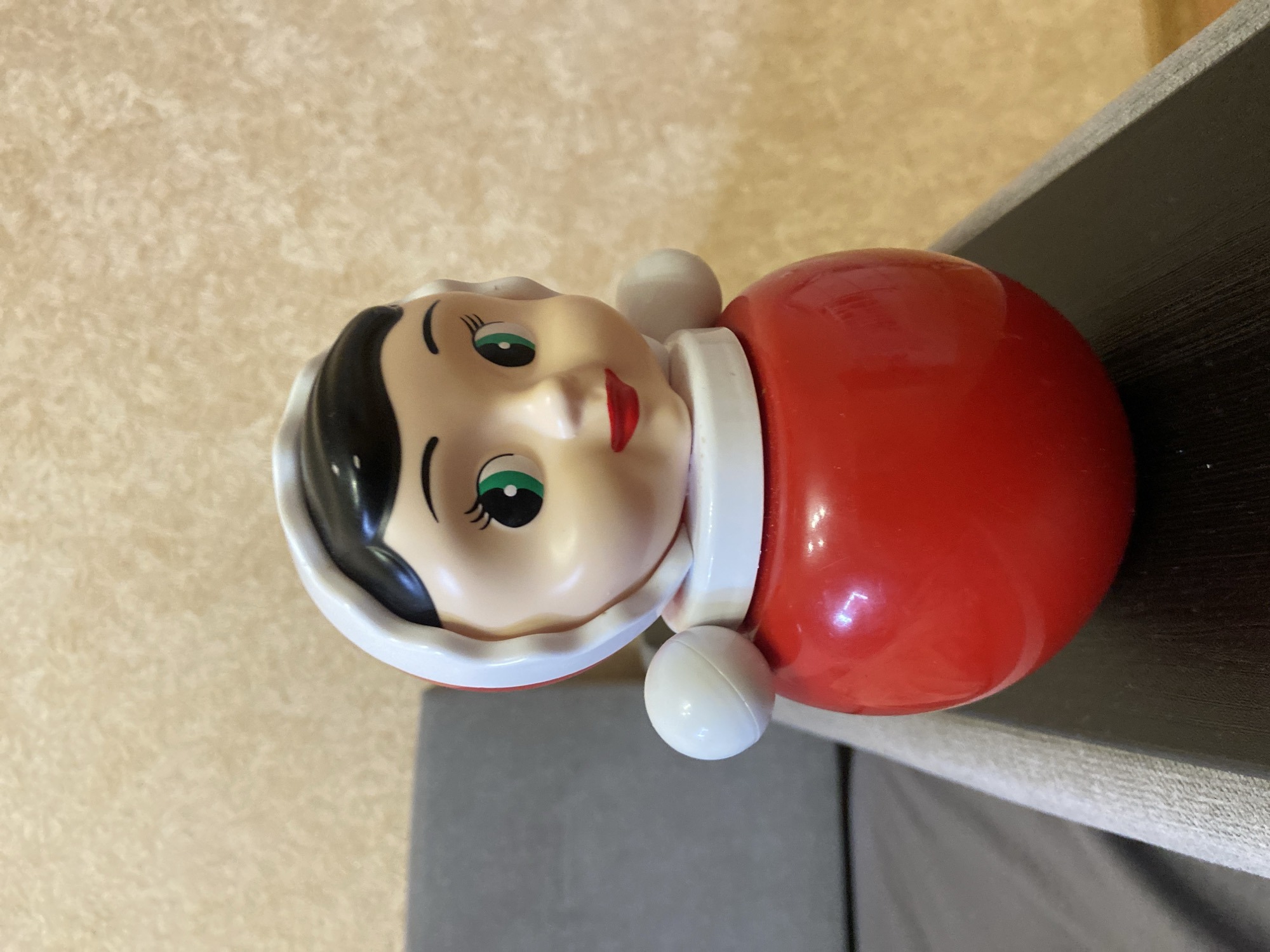 A plastic roly-poly doll with a stylized face and red body is shown on a surface. The doll has a fair complexion, black hair, green eyes, and red lips. It's wearing a white collar and head covering with two white spherical protrusions on either side. The doll's body is red and spherical. The surface the doll rests on is a mix of light brown and gray. The doll seems to be placed on furniture, where different areas are covered with brown and gray texture.
While I can describe the image, I cannot confidently provide the location of the city of Kazan, Russia, since this information is not visually available within the photograph.