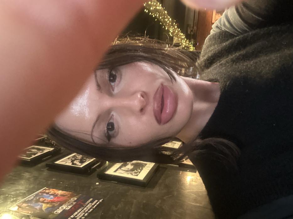 A selfie shows a woman with dark hair and full lips, partially obscured by what appears to be her finger. She wears a dark sweater. Below her are several framed photos and a leaflet titled "Giorgio Scala." String lights are visible in the background. The image is taken at an odd angle, with the woman tilted towards the viewer.