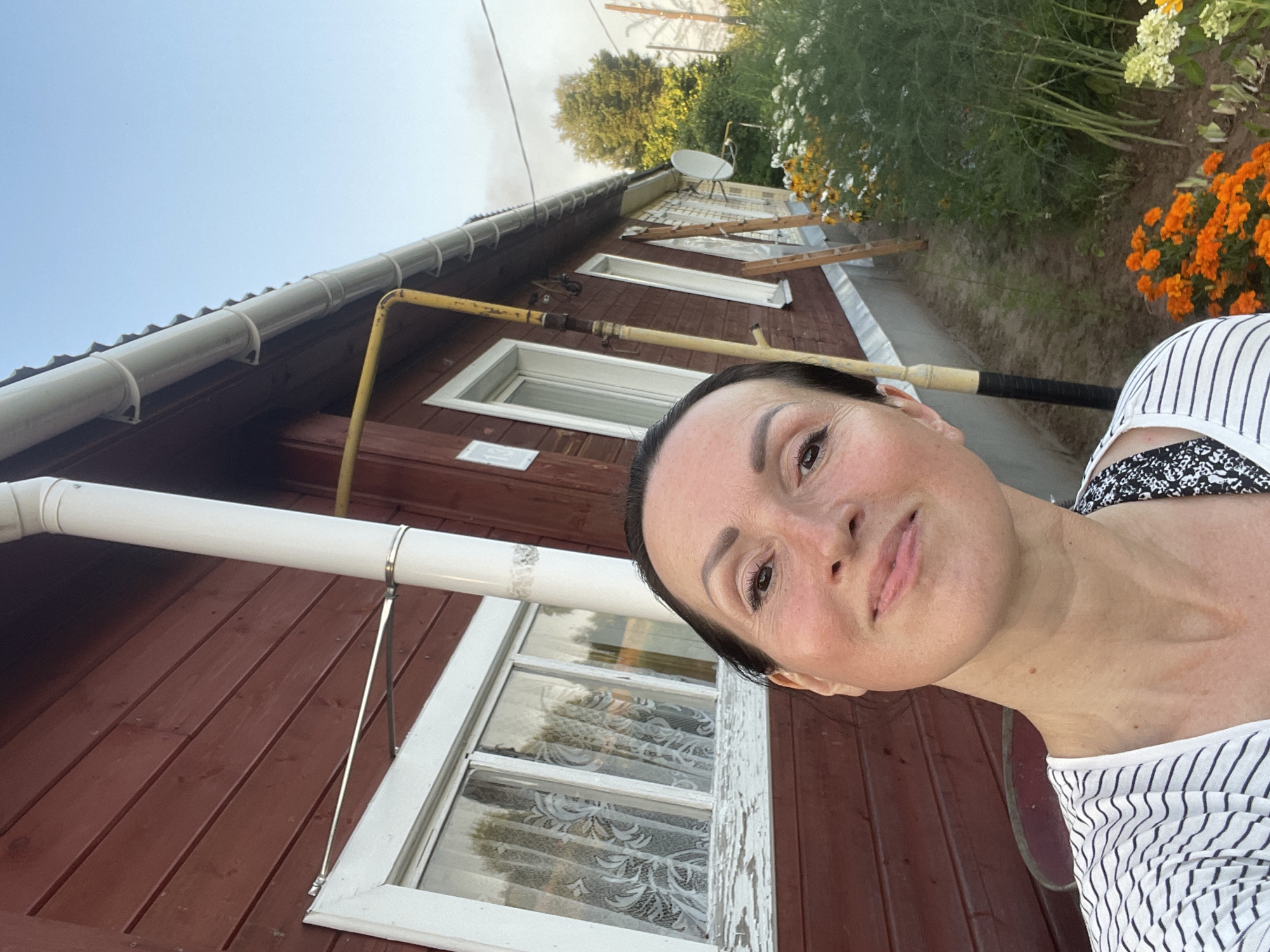 A selfie taken outdoors in the village of Дресвяново, Russia, features a middle-aged woman in the foreground. She has dark hair pulled back, is wearing a striped t-shirt, and has a slight smile. Behind her, a traditional wooden house with red siding and white trim is visible. A white downspout runs vertically along the side of the house, and a yellow gas pipe is also attached to the exterior. A window with white lace curtains is to the left of the downspout. In the background, there are flowers, bushes, and a clear blue sky. The overall scene suggests a residential setting in a rural area.