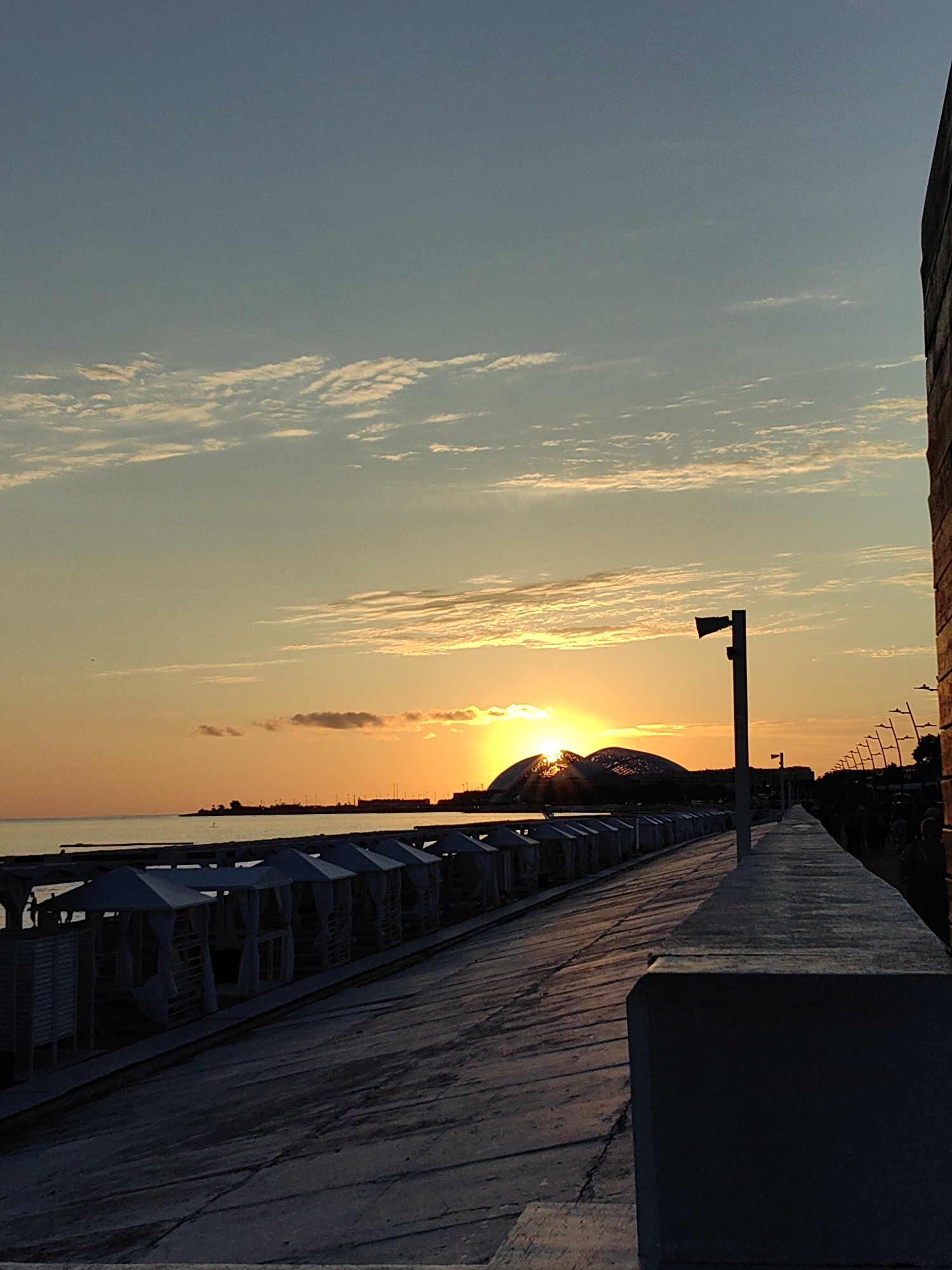 A photograph captures a sunset over the sea in Sirius, Russia. The sky is a gradient of blues and oranges, with scattered clouds. The sun is low on the horizon, partially obscured by a modern, dome-shaped building. In the foreground, a paved walkway runs parallel to the water, lined with white, tent-like structures. Streetlights stand along the walkway, casting long shadows. The sea is calm, with a faint silhouette of land in the distance. The overall atmosphere is serene and picturesque.