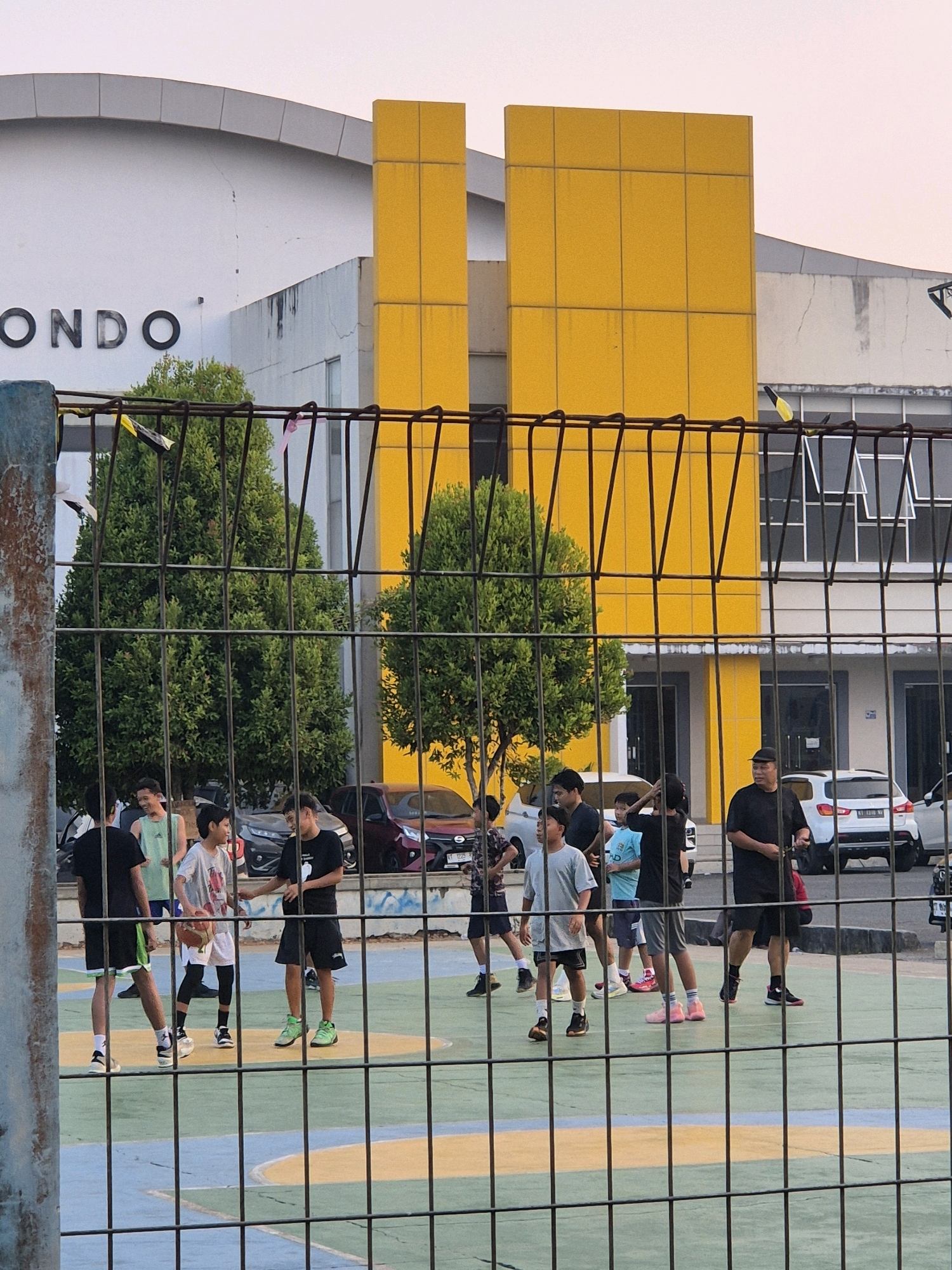 The image depicts a group of young males playing basketball on an outdoor court in Samarinda, Indonesia. The court has a multi-colored surface, with green and yellow markings. In the background, there is a modern building with a distinctive yellow facade and a white section featuring the word "ONDO" in large, dark letters. Several parked cars are visible behind the court, and trees flank the building. The scene is partially obscured by a black metal fence in the foreground. The lighting suggests it is late afternoon or early evening.
