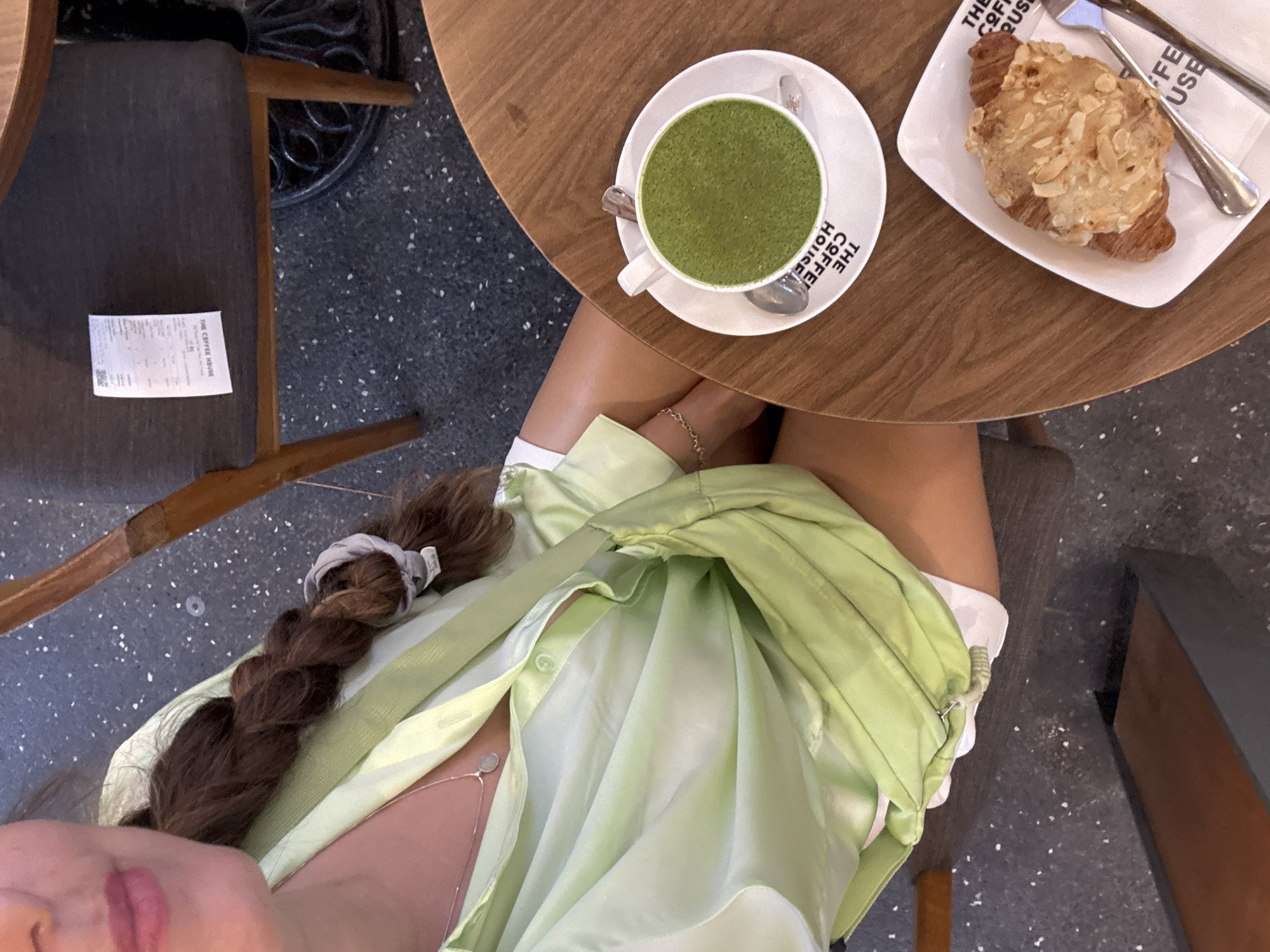 Viewed from overhead, the image shows a woman at a cafe. Her face is partially visible at the bottom left; she has a braid, and is wearing a light green shirt. On the table, there is a cup of matcha latte, a pastry covered in almonds, a fork and a napkin; both items are branded with "The Coffee House." The table is round and made of wood. The cafe has a gray tiled floor with white specks. There is a chair and a receipt on the left. Based on the branding, this image was taken in Nha Trang, Vietnam.