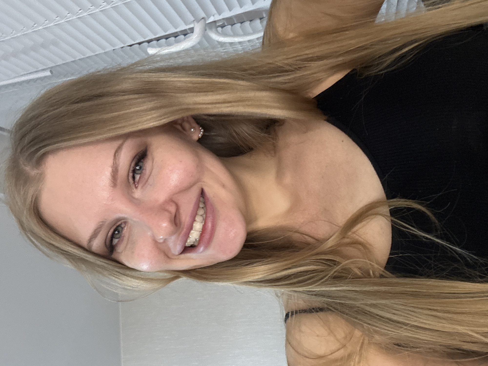 A woman with long blonde hair is smiling at the camera. She is wearing a black top. The background is a white wall and a slatted blind. The location is unknown and the image provides no information to determine if it was taken in Perm, Russia.