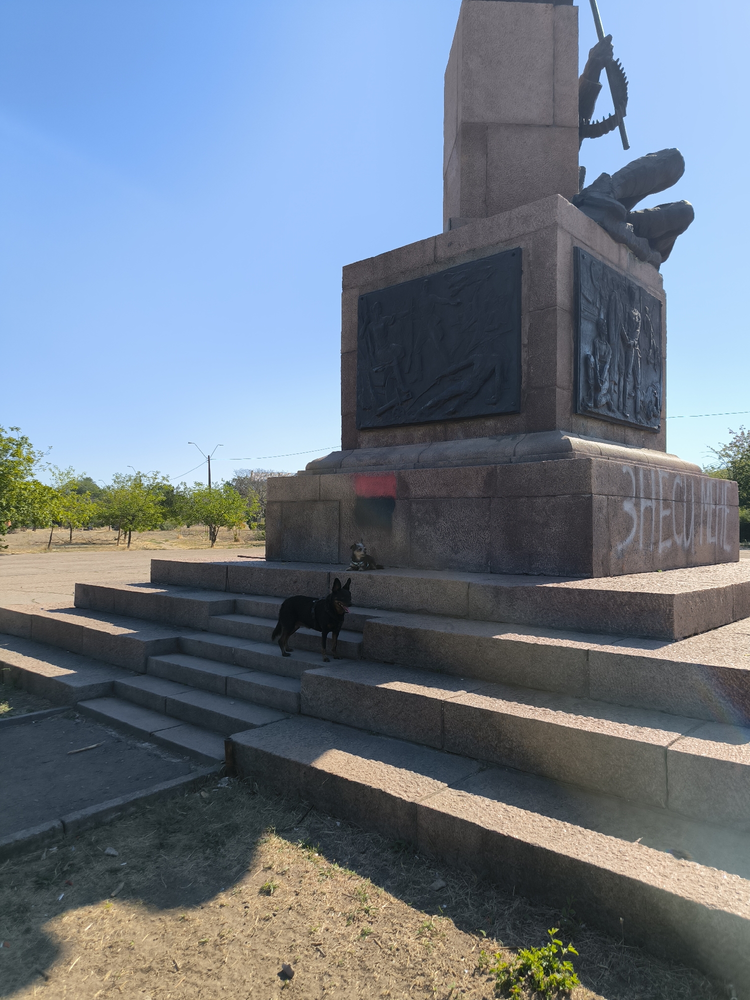 The image shows a monument in the city of Mykolaiv, Ukraine, under a clear blue sky. A medium-sized dark-colored dog stands on the stone steps leading up to the monument. A smaller, lighter-colored dog is also visible resting on the steps. The monument itself is made of stone with large bas-relief panels on its sides and a sculpture at its apex. There is graffiti on the base of the monument, including red and black markings, and white spray paint. In the background, trees and a street light are visible. The area around the monument appears to be a public space with paved and grassy areas.