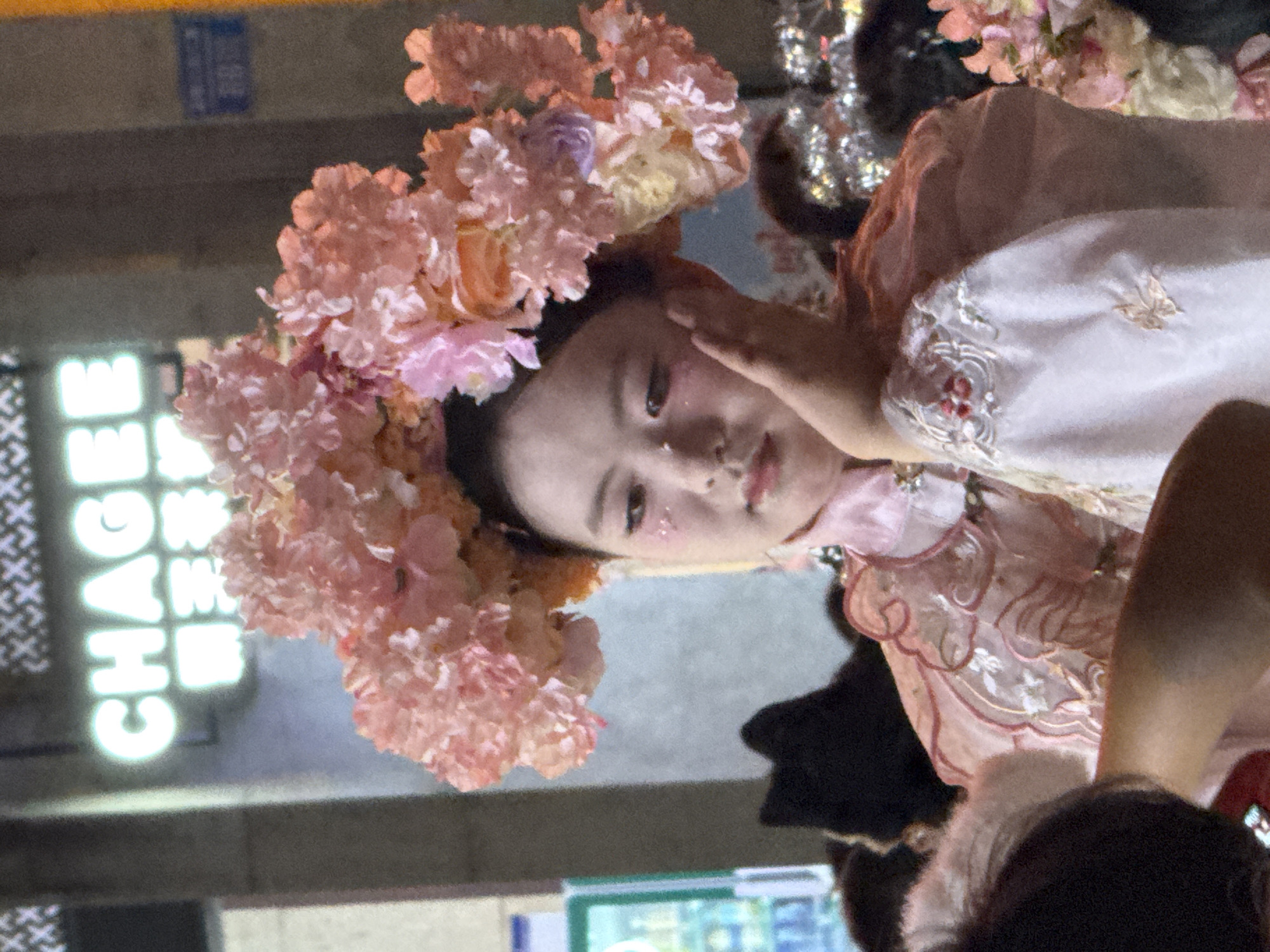A young East Asian female, appearing to be in her late teens or early twenties, is the central subject of the image. She is adorned with a large, elaborate headdress composed of pink and peach-colored artificial flowers. Her hair is dark and pulled back, and she has light makeup on, including what appears to be glitter on her cheeks. She is wearing a traditional-style garment, possibly a qipao or hanfu, in a light pink color with embroidered details and delicate sheer fabric on the sleeves. Her left hand is raised to her face, resting gently on her cheek, while her right arm is lowered. The background is dimly lit, suggesting it is nighttime. A bright, neon sign that reads "CHAGEE" is visible in the upper left quadrant of the image, with Chinese characters below it. The environment appears to be an urban setting, possibly a street or outdoor venue. Other people are present but mostly out of focus in the foreground and background. The lighting is somewhat harsh, with a focus on the subject. The overall impression is of a cultural event or performance. The location is Yuzhong District, China.