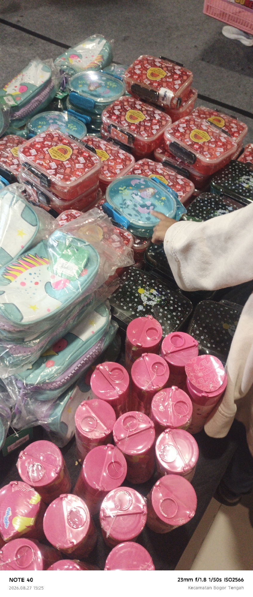 The image shows a close-up, overhead view of several children's lunchboxes and containers, primarily in pink and turquoise, stacked and displayed on a dark surface. Some items feature cartoonish designs, including unicorns and cartoon characters. In the foreground, a hand, likely belonging to a child, is visible, pointing at one of the turquoise lunchboxes. In the lower right corner, a text overlay indicates the location "Kecamatan Bogor Tengah, Bogor, Indonesia" and camera settings. The overall scene appears to be an informal market or retail display.