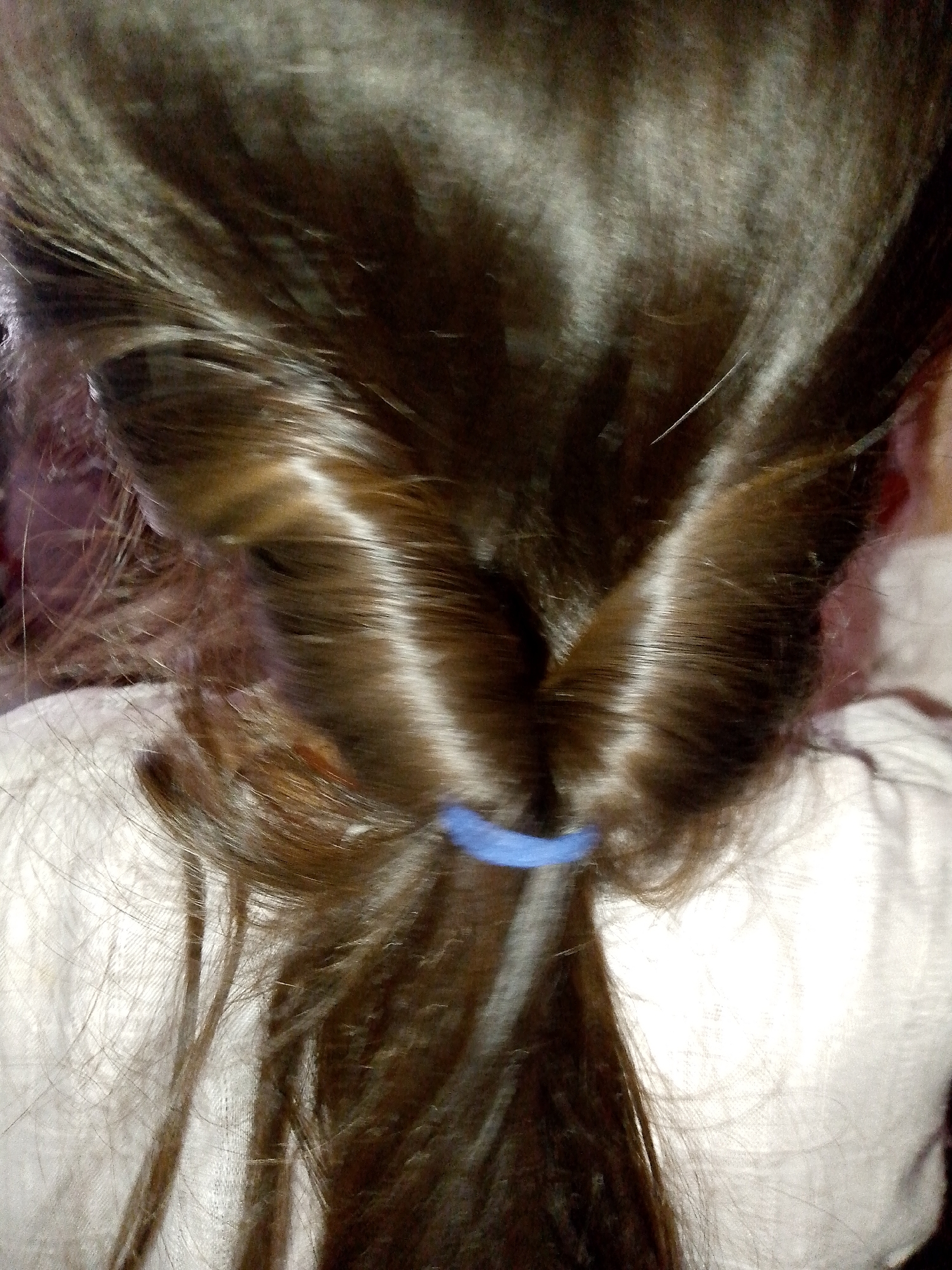 The image is a close-up, top-down view of a person's dark brown hair styled in a braid, held in place with a blue hair tie. The hair is shiny, suggesting good health or product use. The background is blurred and appears to be white fabric, possibly clothing or bedding. The lighting highlights the texture and individual strands of the hair, creating streaks of light and shadow. There are no visible features of the person's face or body, nor any context related to a specific city or location.