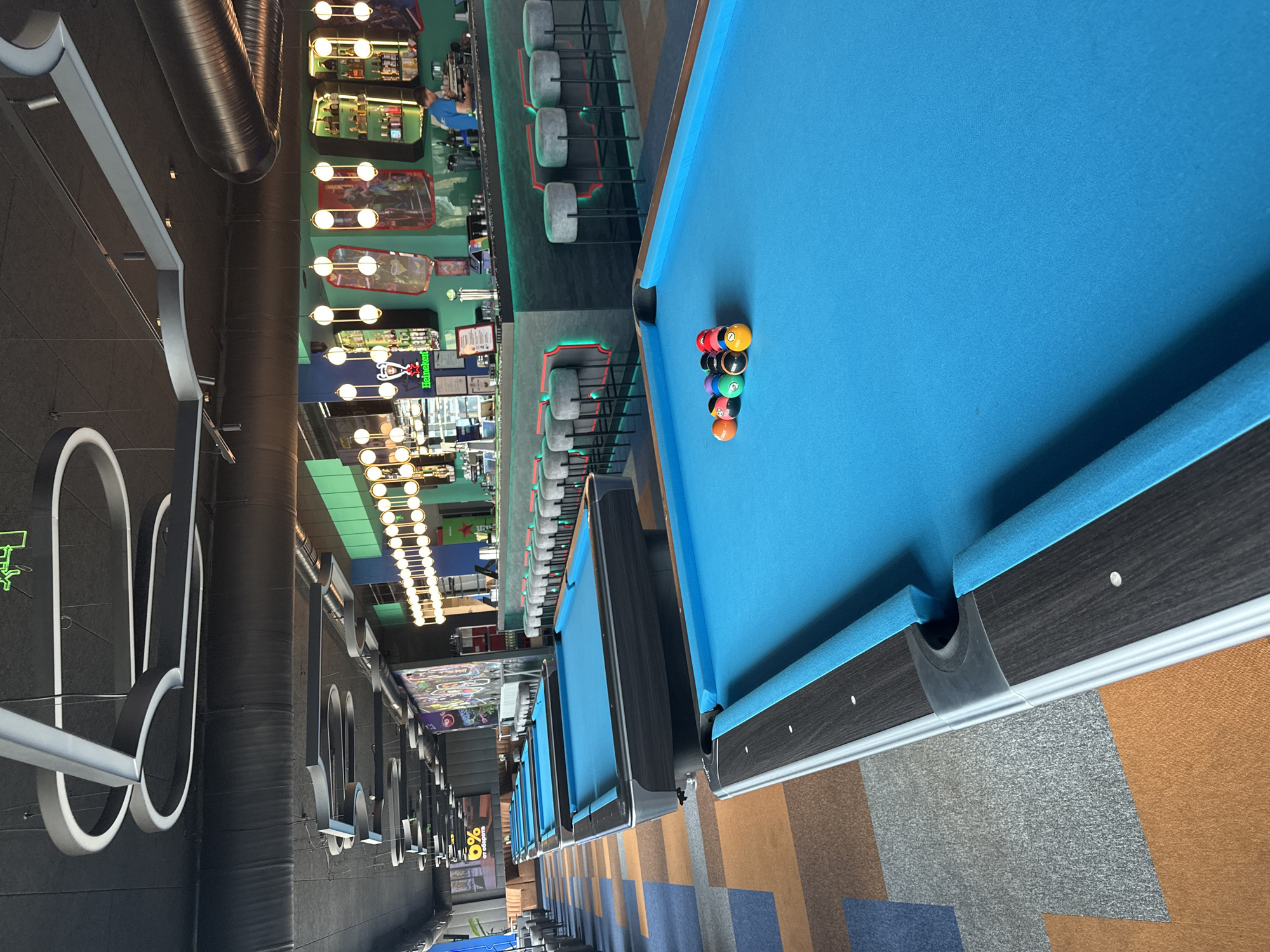 The image shows the interior of a modern billiards hall located in Varna, Bulgaria. The hall features multiple blue-felt pool tables, arranged in rows, with one table prominently in the foreground. On this table, a rack of billiard balls is set up, consisting of solid-colored balls and striped balls, as well as the cue ball. The environment is brightly lit by overhead track lighting with a sleek, contemporary design. In the background, a bar area is visible, complete with stools, bottles of liquor, and a bartender serving customers. The floor is covered in a colorful, patterned carpet with geometric shapes in shades of blue, orange, and brown. The walls are decorated with vibrant artwork and promotional posters, contributing to the lively atmosphere of the establishment. The overall scene depicts a well-maintained and inviting entertainment venue.