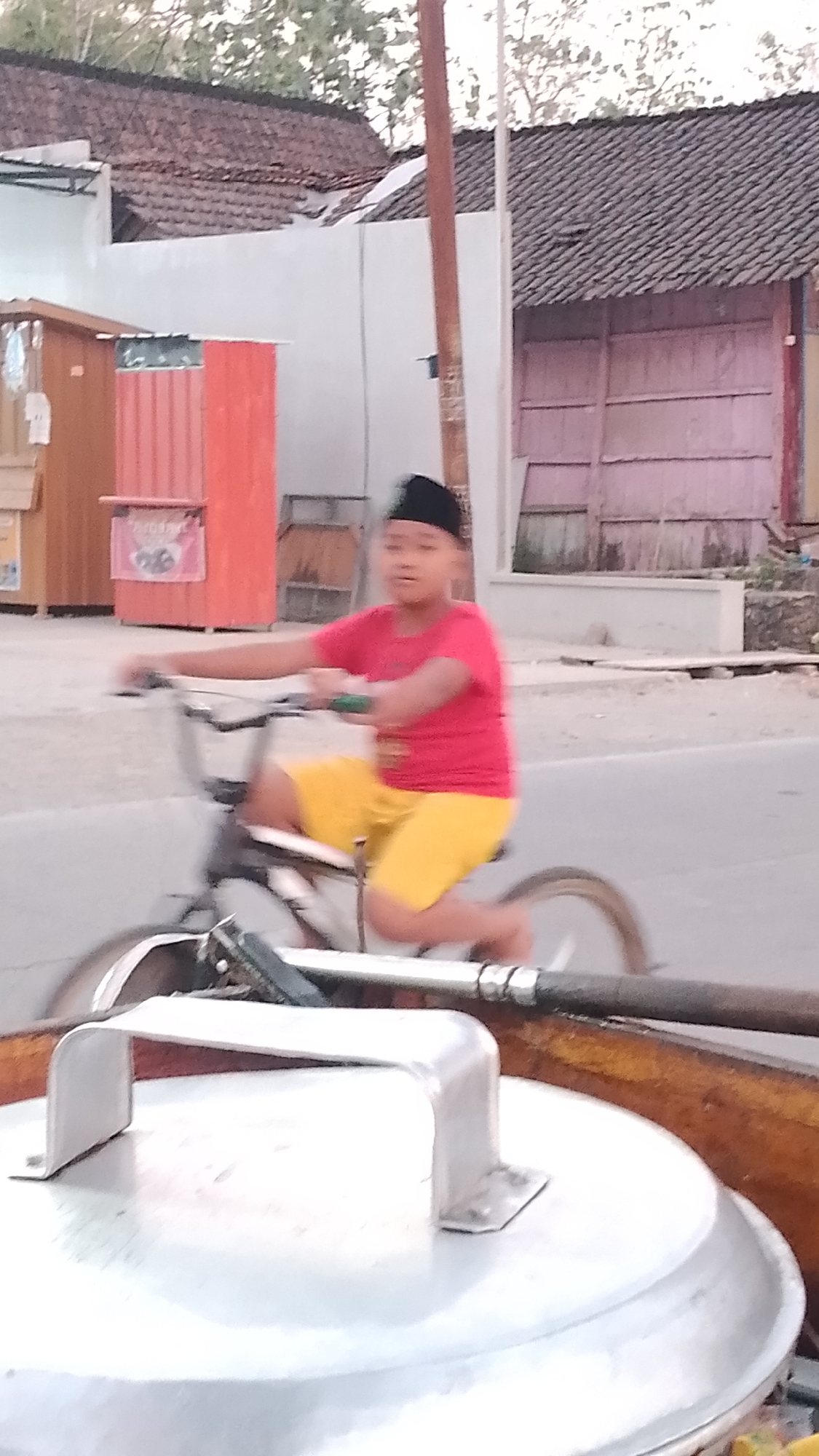 In Karanglangu, Indonesia, a young boy wearing a black peci and a red t-shirt is riding a bicycle. He is also wearing yellow shorts. The boy is in motion, with his right arm extended forward and his left arm bent. The bicycle has a black frame and appears to be a BMX style bike. The background shows a street with buildings and trees. One building has a red corrugated metal wall, and another has a pink wooden wall. There is also a wooden pole in the foreground. In the immediate foreground, a large, silver pot lid with a silver handle is visible. The lighting suggests it is either early morning or late afternoon.