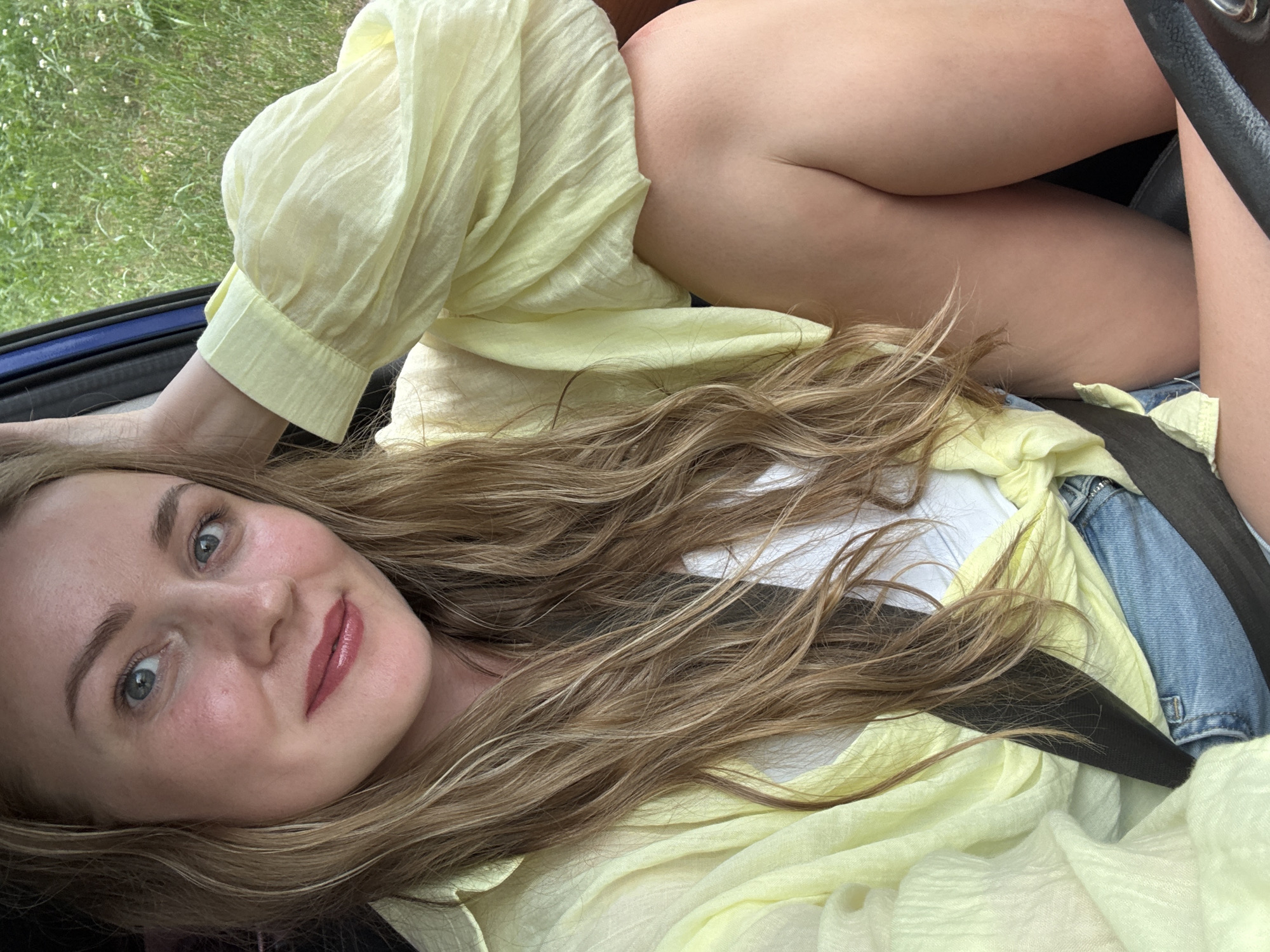 A young woman with blonde hair and blue eyes is sitting in a car, her arm resting on her head. She is wearing a yellow blouse and denim shorts. Her body is angled towards the right side of the frame. The background shows green grass and some foliage, suggesting an outdoor setting. The image captures a candid moment, likely taken during a car ride.