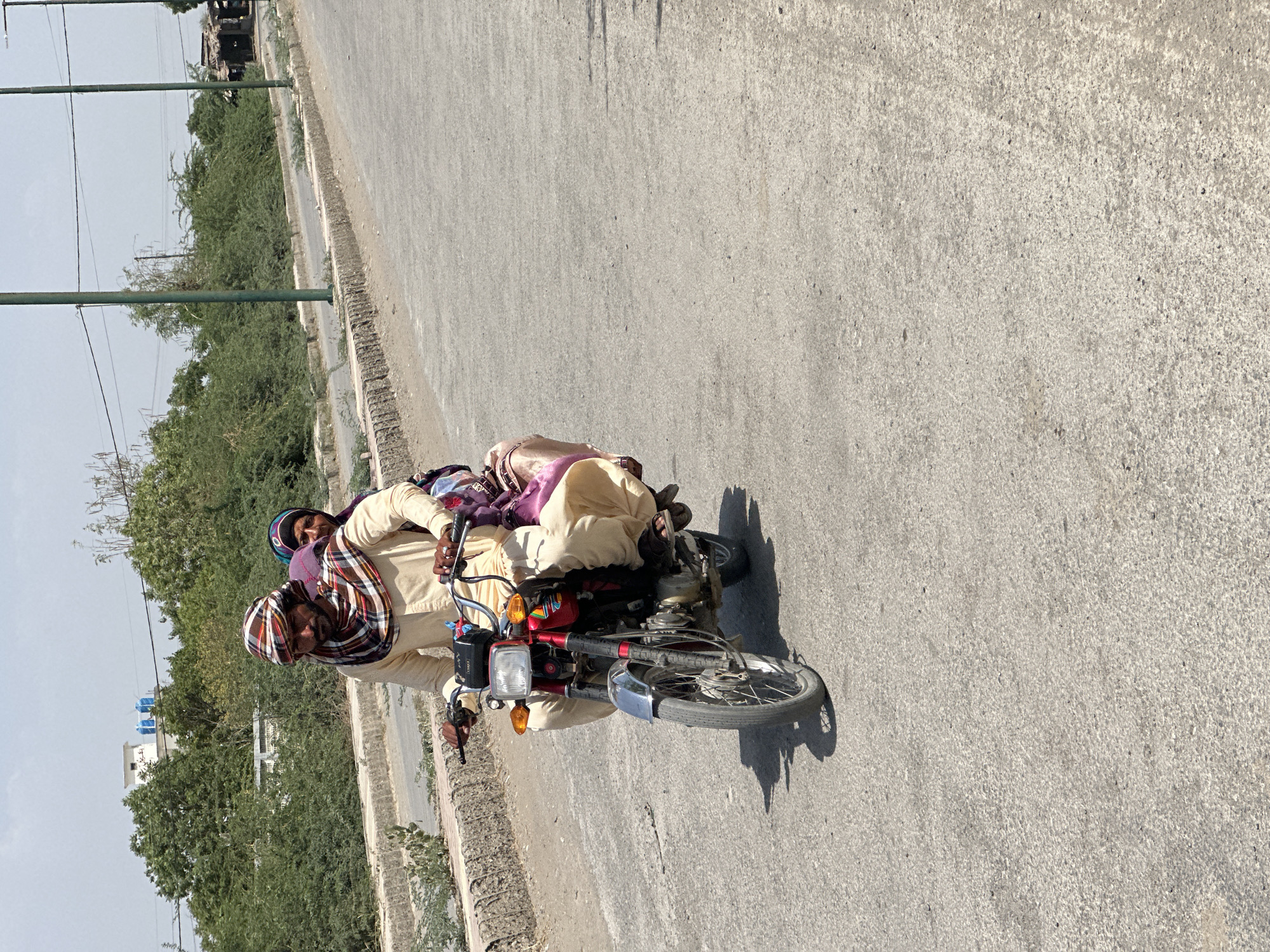 A man is riding a red and blue motorcycle on a concrete road. He has a patterned scarf wrapped around his head and is wearing a light-colored shirt. A second person is sitting behind him on the motorcycle. Green trees line the road in the background, and some buildings are visible in the distance. The location is Unknown, Unknown.