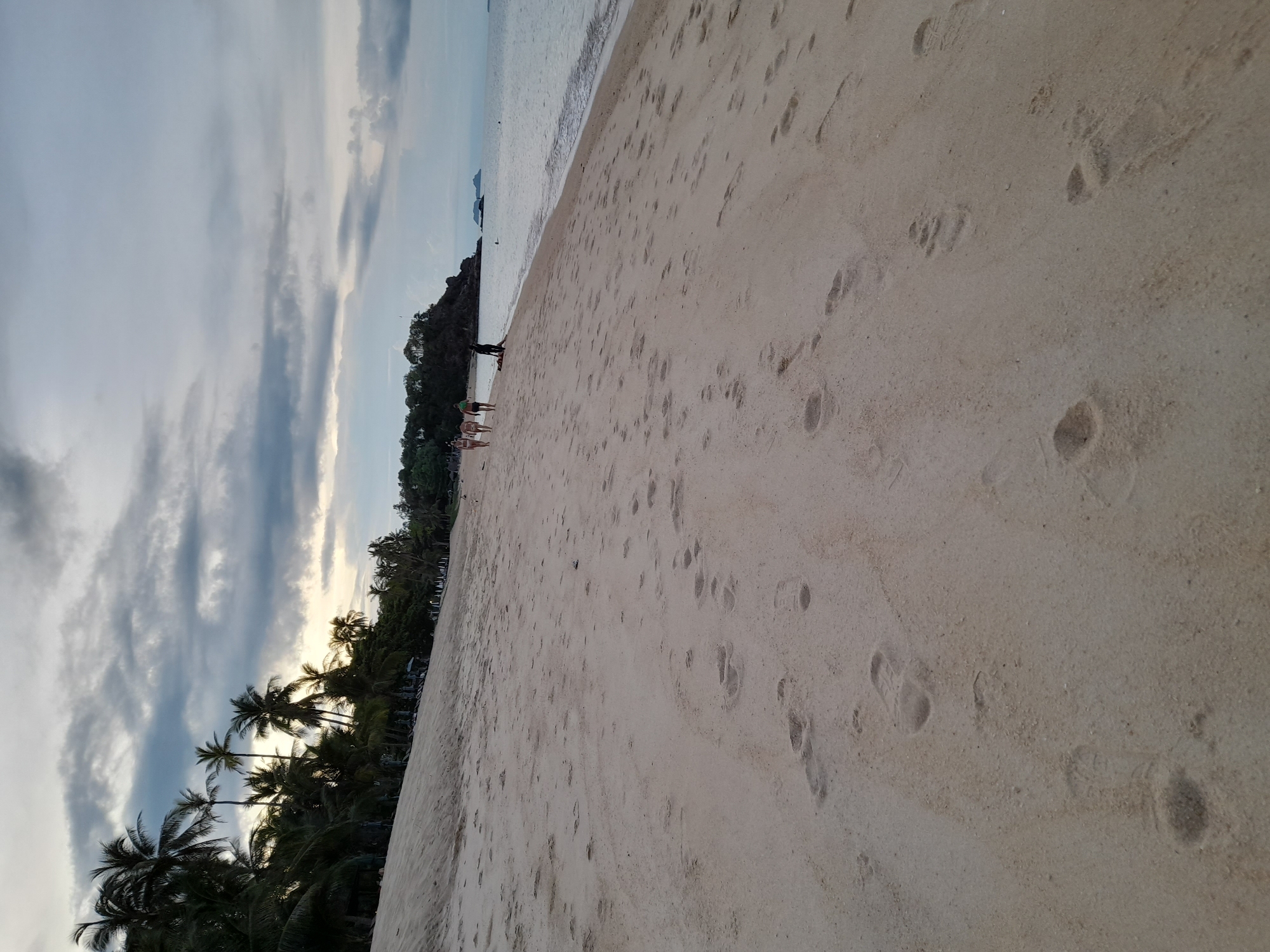 The image depicts a wide sandy beach in Baan Tai, Thailand, at dusk. The tide is low, revealing a vast expanse of wet sand imprinted with numerous footprints, some of which appear to be from shoes and others from bare feet. Along the left side of the beach, a dense line of palm trees and other tropical vegetation creates a natural border, with a few lounge chairs visible amongst the trees, suggesting a resort or public beach area.
In the middle ground of the scene, a small group of four individuals is walking along the shoreline, heading towards the right of the frame. Two of the individuals are closer to the water, while the other two are further up on the sand. A fifth individual is partially visible lying down on the sand closer to the water, with their legs extended towards the sea. In the distance, on the right side of the image, the calm ocean stretches out to the horizon, meeting a cloudy sky. Two small islands or rocky outcrops are visible in the water further offshore, adding depth to the seascape. The overall atmosphere is serene and tranquil, characteristic of a tropical evening.