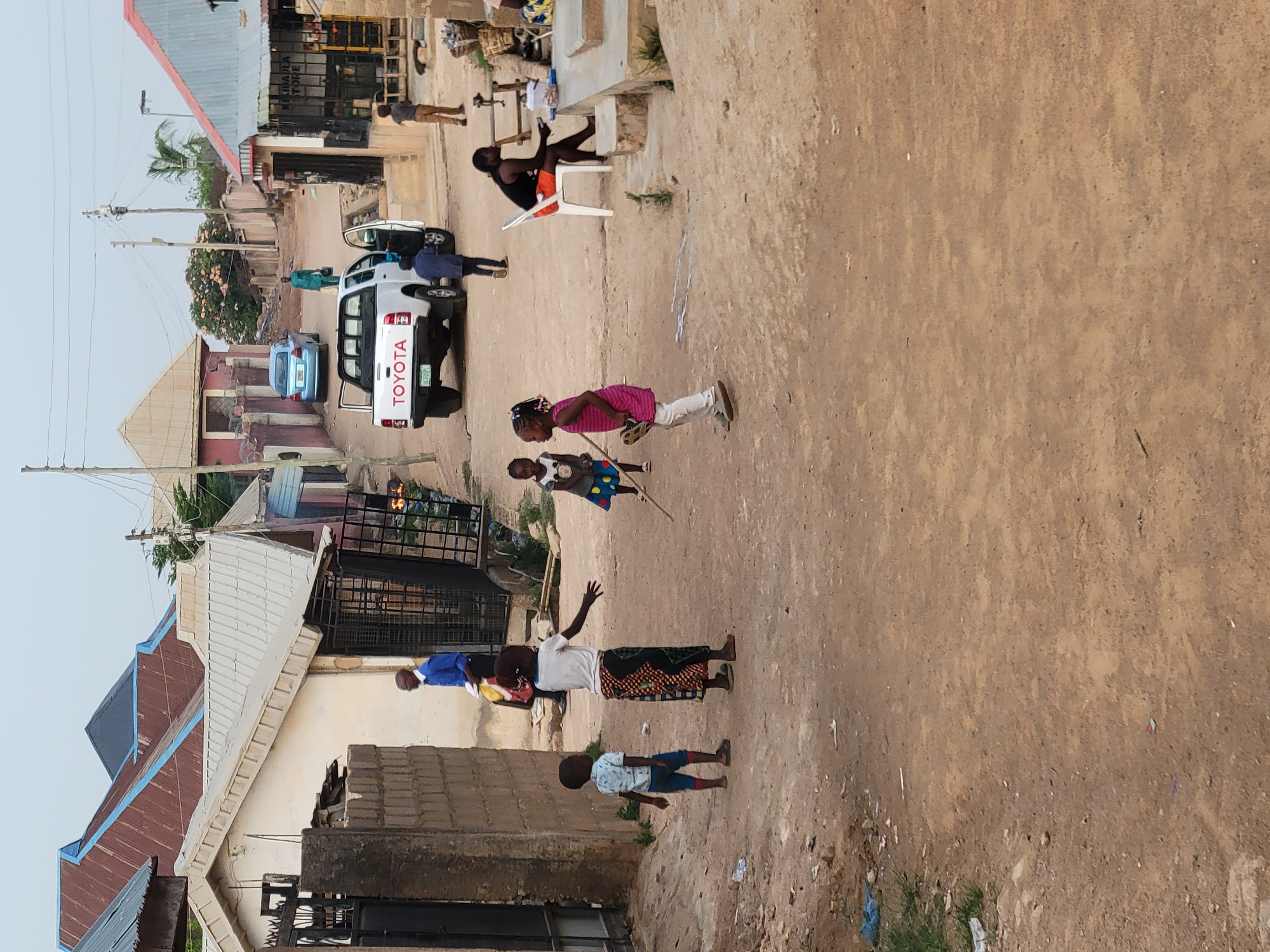 The image depicts a street scene in what appears to be Jos, Nigeria. The road is unpaved and dusty. Several people are present, including children playing in the road. One child holds a stick. A white Toyota pickup truck is parked on the side of the road. Houses and buildings line the street. A man is seated on a white plastic chair outside one of the buildings. The sky is overcast.