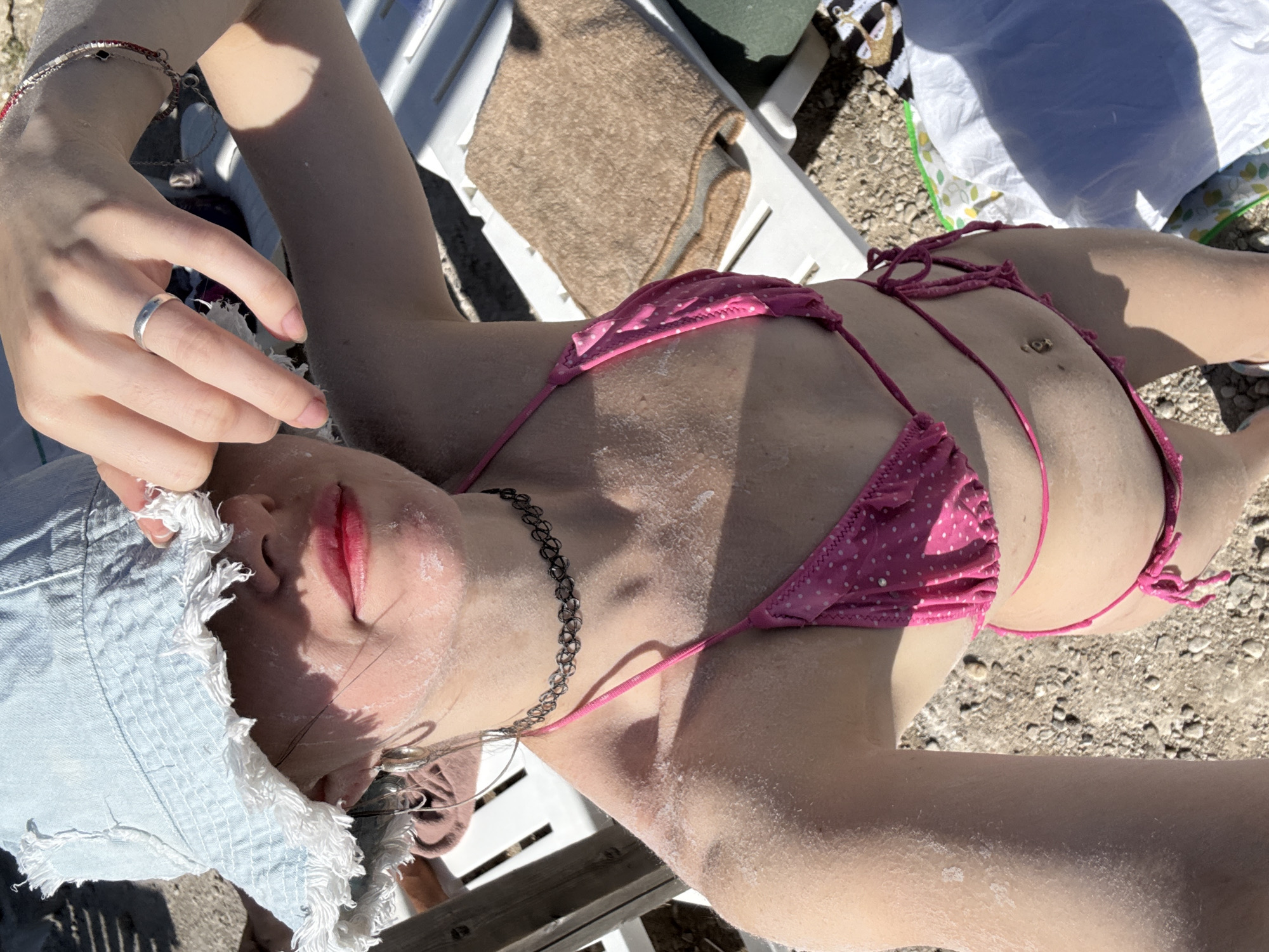 A person wearing a light blue denim bucket hat and a pink polka-dot bikini is lying on a white chaise lounge outdoors. The person's face is partially obscured by their hand, which is resting on the brim of the hat. There appears to be white residue, possibly sunscreen, on their face and shoulders. A silver ring is visible on the person's left ring finger, and a thin black choker necklace is around their neck. The background suggests an outdoor setting with sandy ground and some vegetation. The overall impression is of someone enjoying a sunny day, likely at a beach or resort. The image is cropped from the chest up, with a portion of the bikini bottom and belly button visible.
