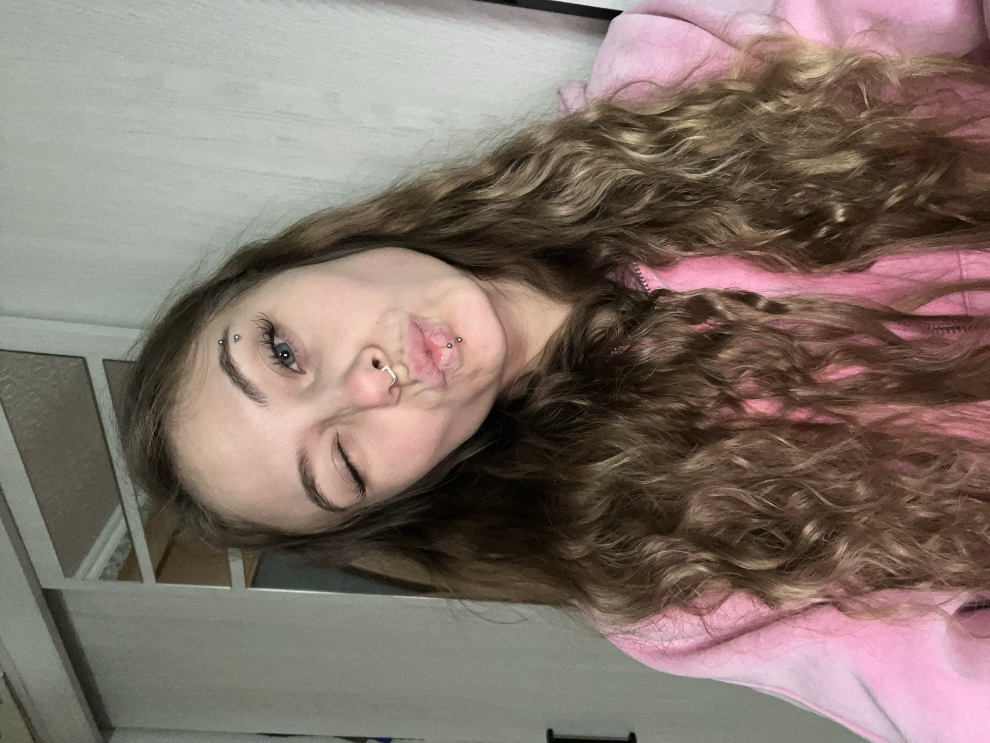 A young woman with long, curly brown hair, a nose ring, an eyebrow piercing, and lip piercings is wearing a pink hoodie. She is puckering her lips and winking. The background includes part of a white painted wooden frame or headboard and a white wall. There is no indication of location within Фоки, Russia.