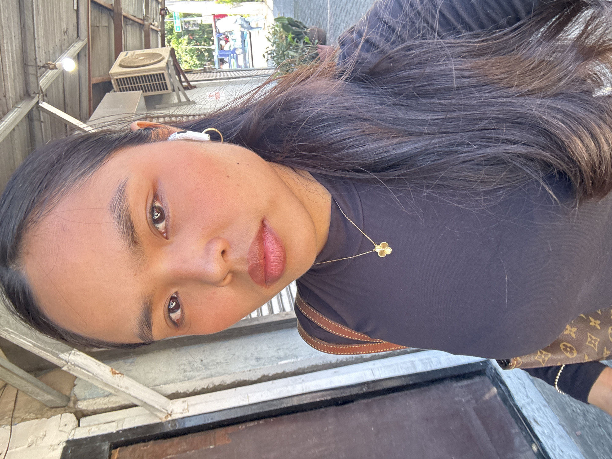 A young woman with dark hair and brown eyes is taking a selfie outdoors. She is wearing a black shirt, a gold clover necklace, and a white earbud. A brown Louis Vuitton purse is slung over her shoulder. In the background, there is a building with an air conditioning unit and some plants. The scene appears to be on a street in Davao City, Philippines, possibly near a market or commercial area, given the presence of shops and the general urban ambiance.