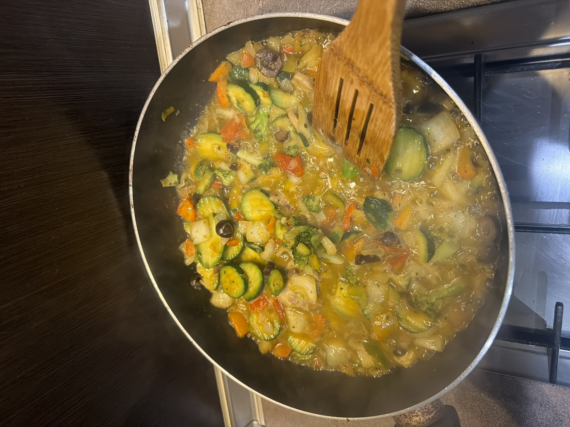 A top-down, eye-level shot captures a black frying pan filled with a colorful medley of sautéed vegetables, including sliced zucchini, chopped carrots, broccoli florets, and black olives. The vegetables are submerged in a glistening, golden broth, with steam rising from the pan. A wooden spatula with slotted grooves is positioned on the right side of the pan, partially submerged in the food, indicating recent stirring or serving. The pan rests on a stainless steel stovetop with visible grates and burners, suggesting a kitchen environment. The background is a dark, textured surface, possibly a cabinet or backsplash. The scene is set in an indoor kitchen. The city of Ruse, Bulgaria is not depicted or suggested by any elements in the image.