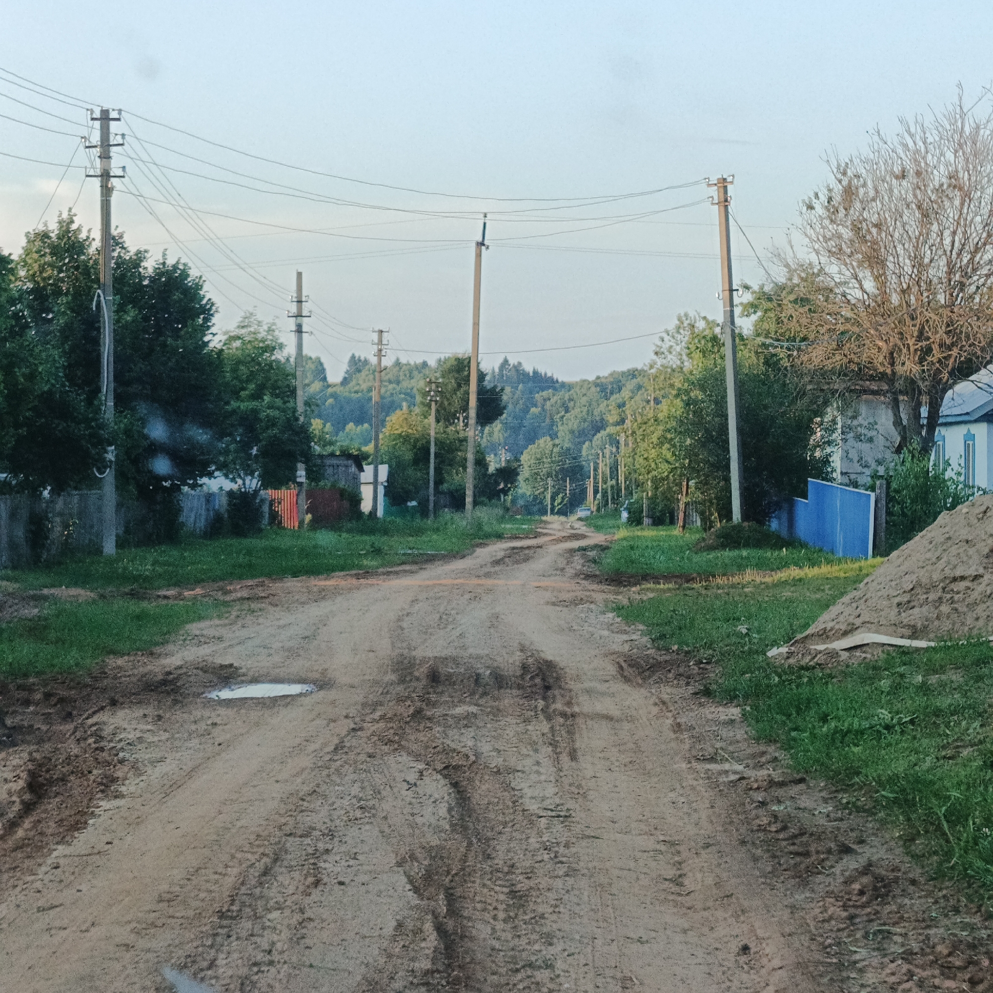 The image depicts a dirt road in the village of Vanky, Russia. The road is unpaved and shows signs of wear and tear, with tire tracks and a puddle visible. On either side of the road, there are trees and grass. In the background, there are houses and power lines. The sky is clear and blue, suggesting it is daytime.