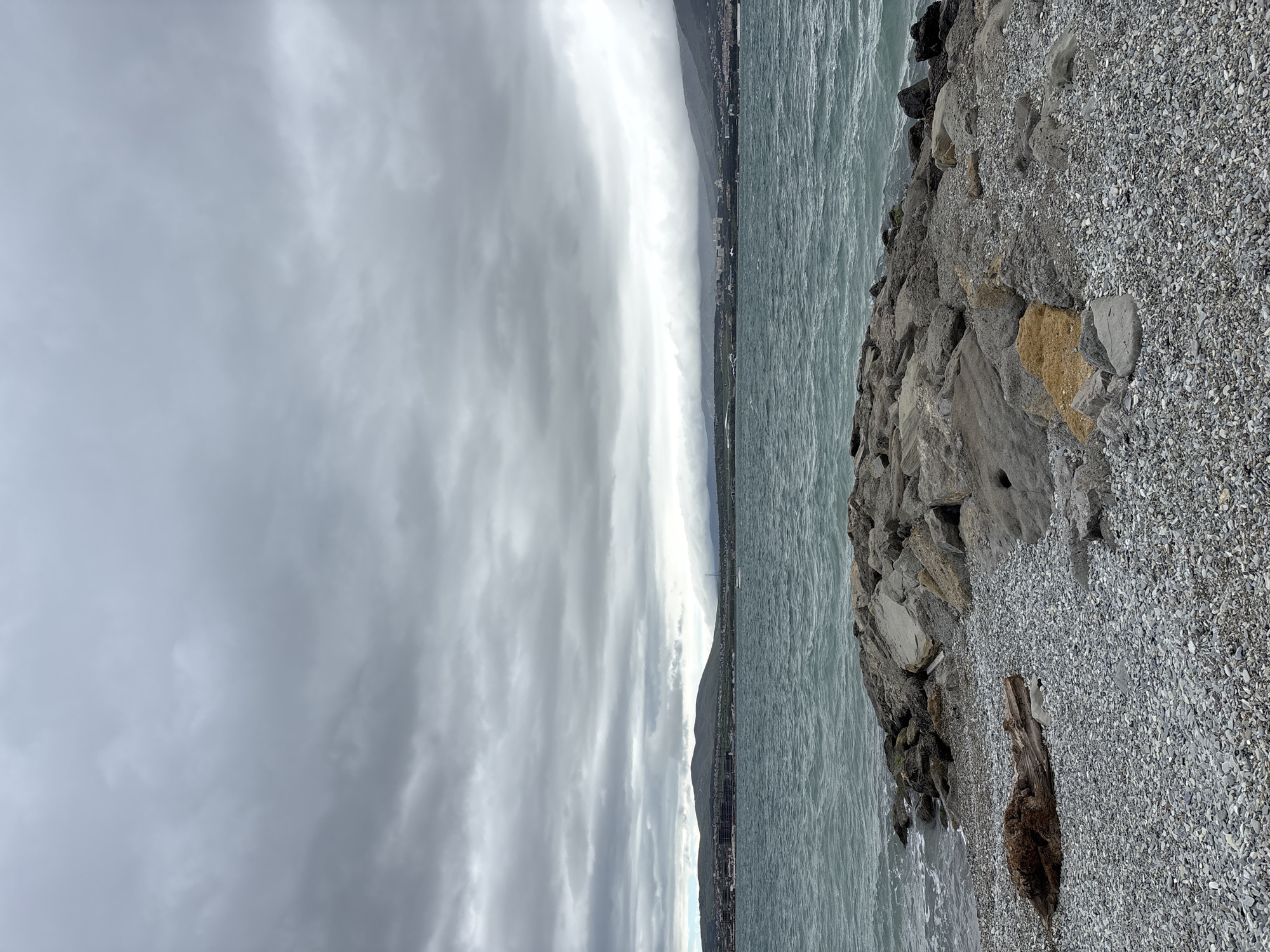 The image captures an overcast seascape, likely near Gelendzhik, Russia. Dominating the foreground is a rocky shoreline transitioning into a pebble beach. The water, a pale gray-green, stretches towards a distant horizon, where a thin strip of land is visible, possibly indicating the city. A thick blanket of gray clouds covers the sky, creating a somber atmosphere. There is a hint of light breaking through the clouds in the center. There are no people visible in the frame. A piece of driftwood rests on the pebble beach. The overall impression is one of a cold and slightly desolate coastal scene.