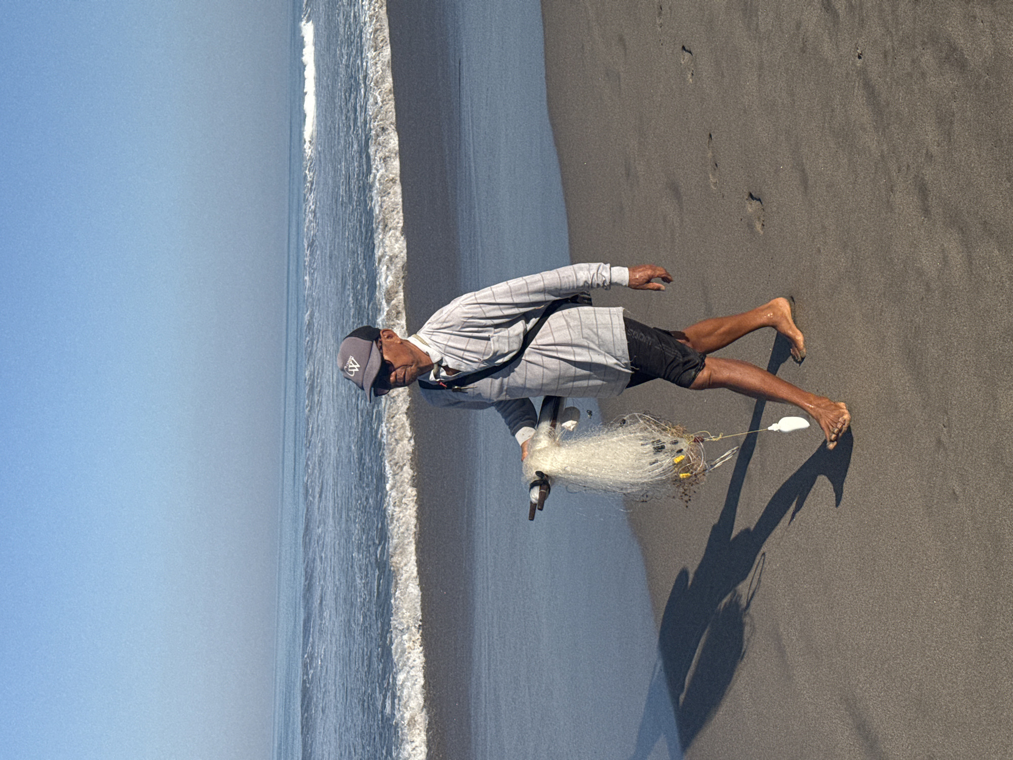 A man stands on a sandy beach, holding a fishing net. He is wearing a gray striped long-sleeved shirt, black shorts, and a gray baseball cap. A strap goes across his torso. The beach stretches out towards the ocean, where waves are visible. The location is North Kuta, Indonesia.