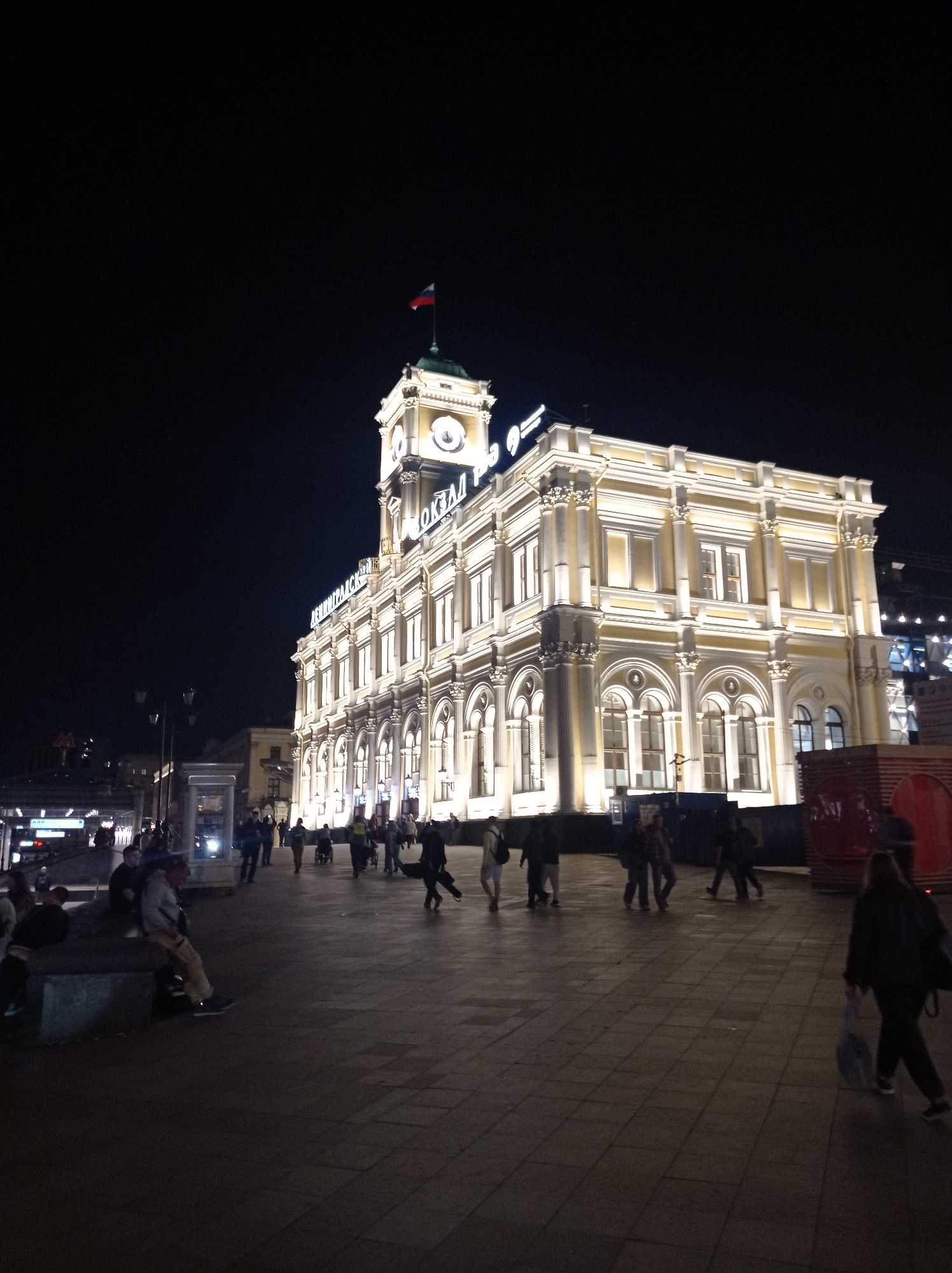 An exterior, eye-level, nighttime photograph captures the brightly lit facade of a large, ornate building in Moscow, Russia. The building, likely a train station given the visible Cyrillic signage, is illuminated with warm yellow lights, highlighting its classical architectural features, including arched windows and columns. A Russian flag flies from a small tower atop the building, which also features a clock face. In the foreground, a paved plaza is populated by a few individuals walking, sitting, and standing. Some are silhouetted against the building's light. The overall atmosphere is that of a well-lit, public urban space at night.