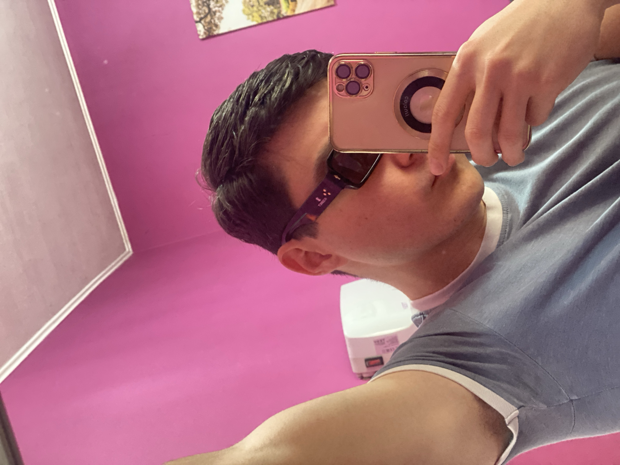 The photograph depicts a person taking a selfie in a room with a pink wall. The person is holding a gold-colored iPhone up to their face, partially obscuring their nose and mouth. They are wearing sunglasses with a dark lens and a frame that appears to be purple or burgundy. They are wearing a blue short-sleeved T-shirt with white trim at the collar and sleeves. Behind the person is a white hand dryer mounted on the pink wall. Above the person is a piece of art or a photo on the wall. The scene appears to be indoors, possibly in a bathroom or dressing room. The location within the city of Varna, Bulgaria, cannot be determined from the image alone.